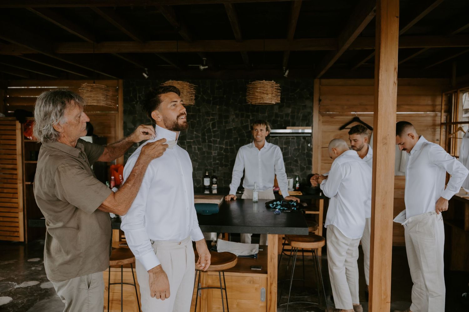 Groomsmen in white linen suits helping each other get dressed before a wedding ceremony.