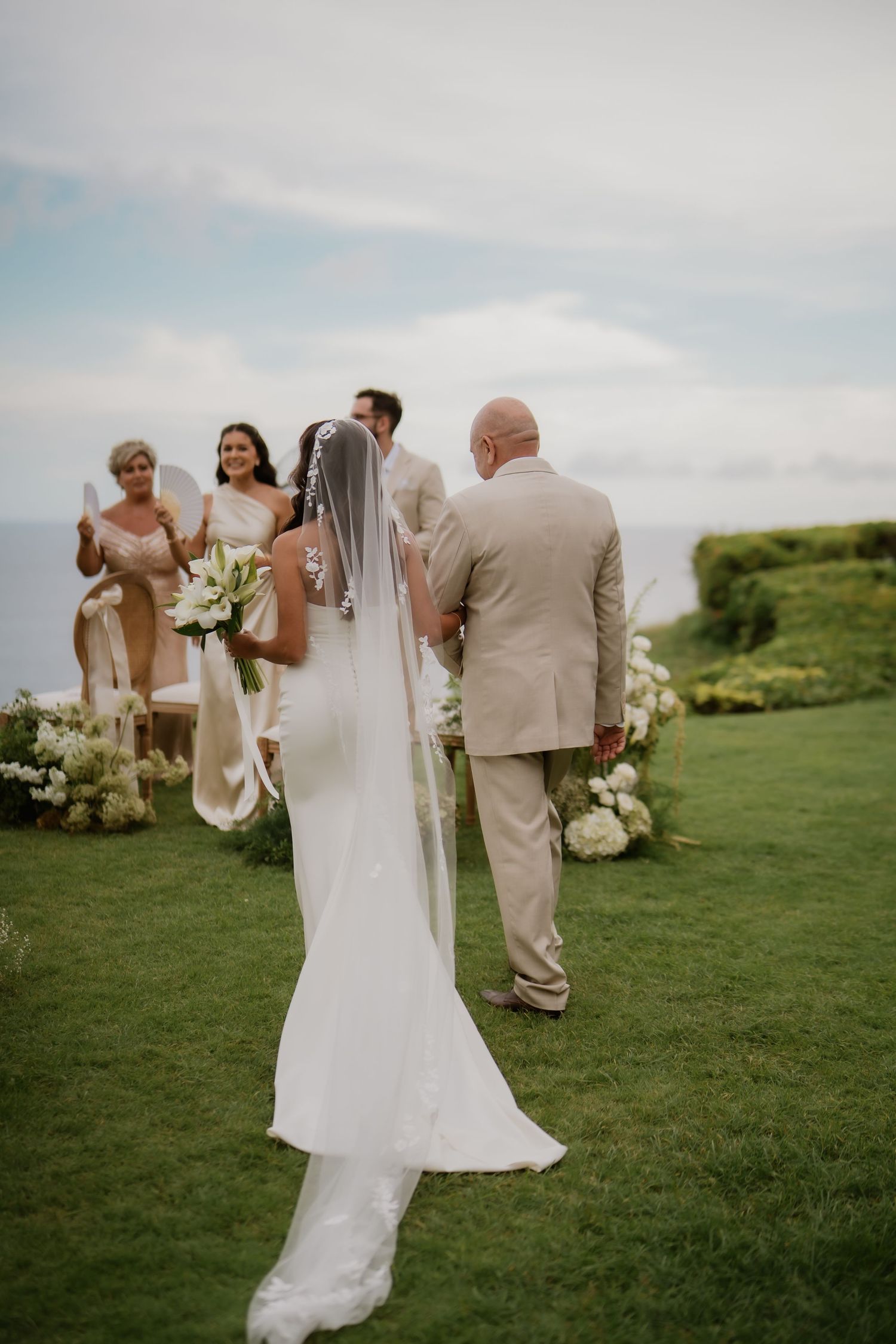 Bride in white gown and veil walking down the aisle with escort toward ocean-view altar.