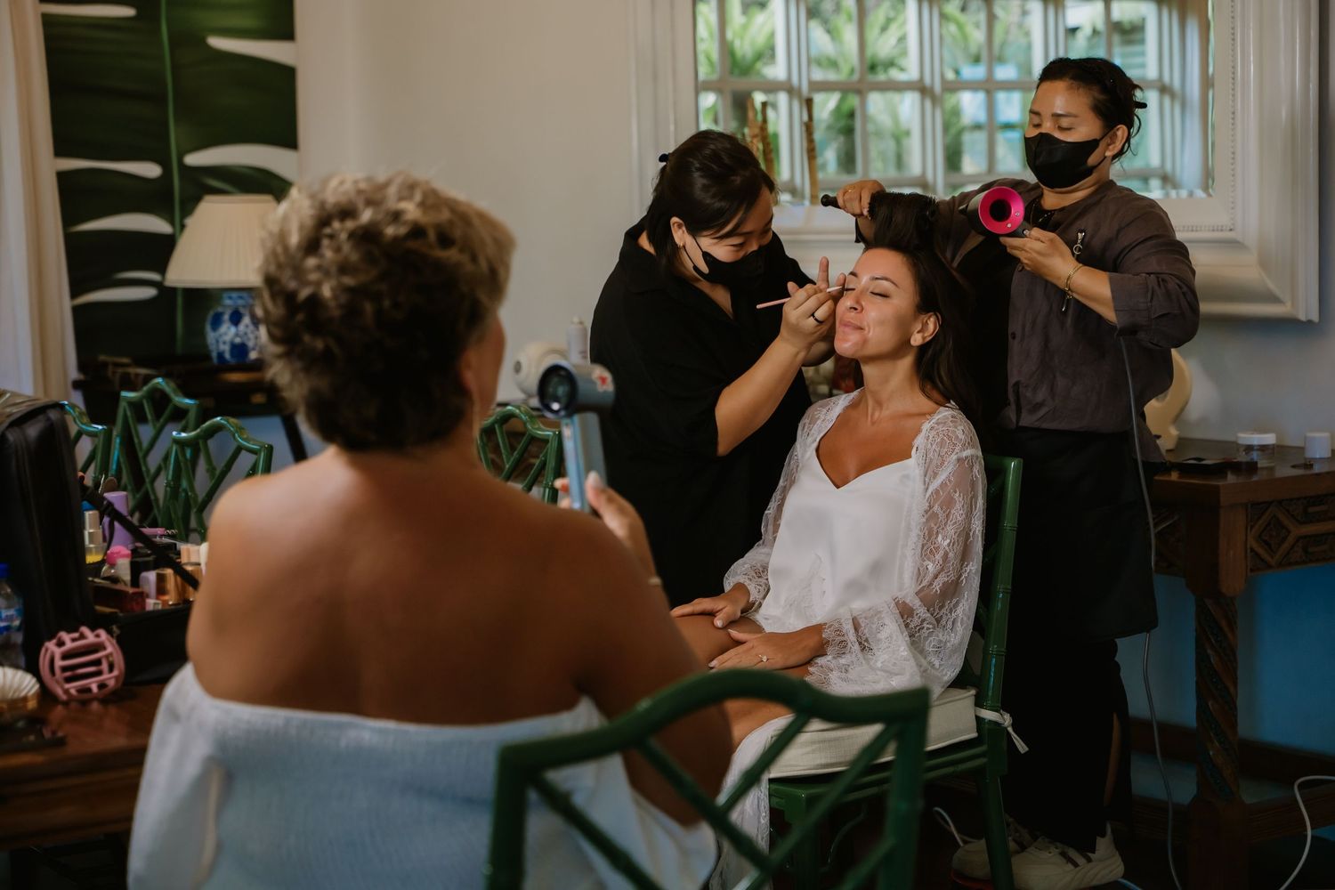 Bride getting hair and makeup done by two stylists wearing masks in a tropical wedding preparation room.