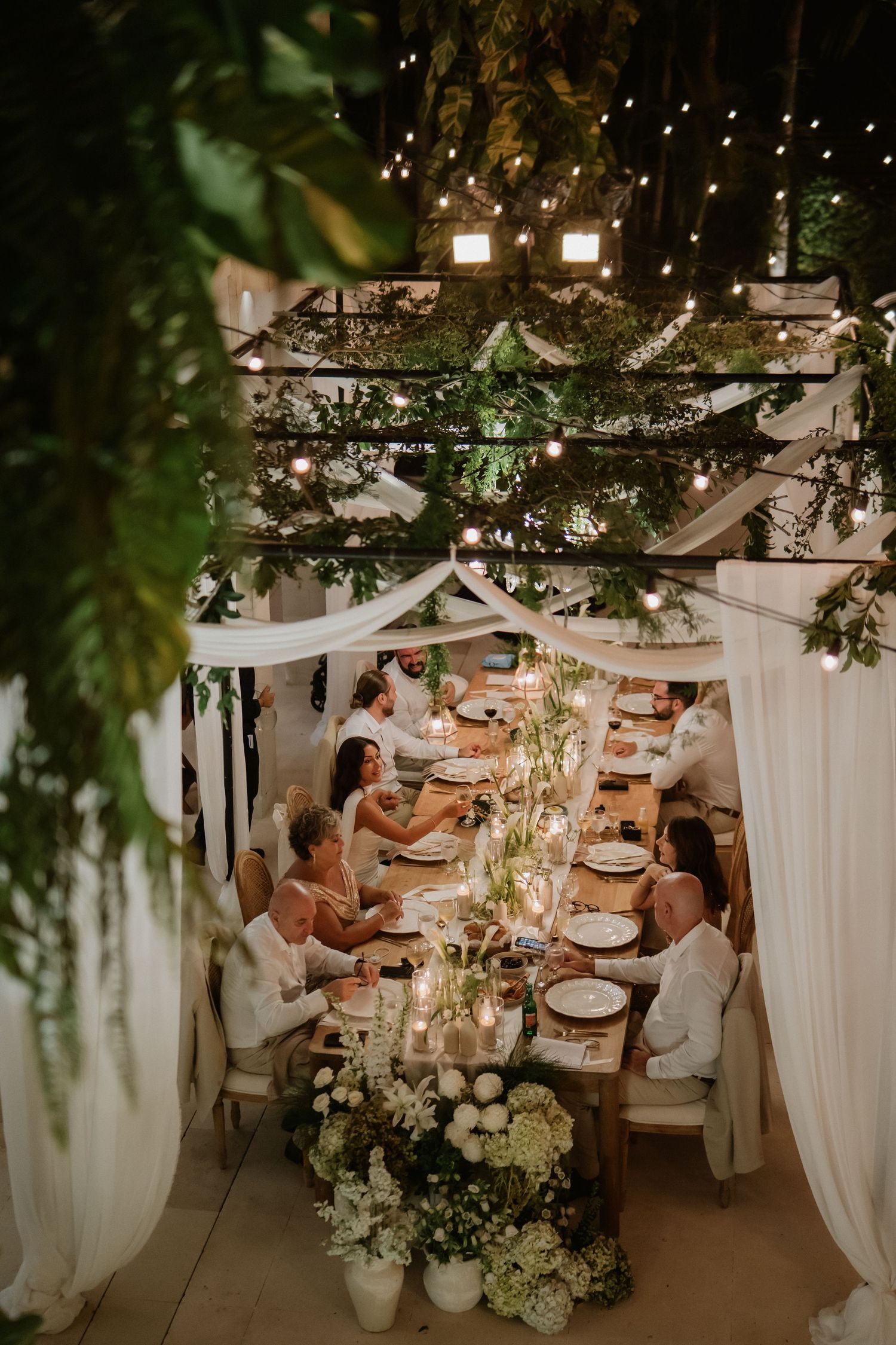 Overhead view of intimate wedding reception dinner under string lights with lush greenery and white floral centerpieces.