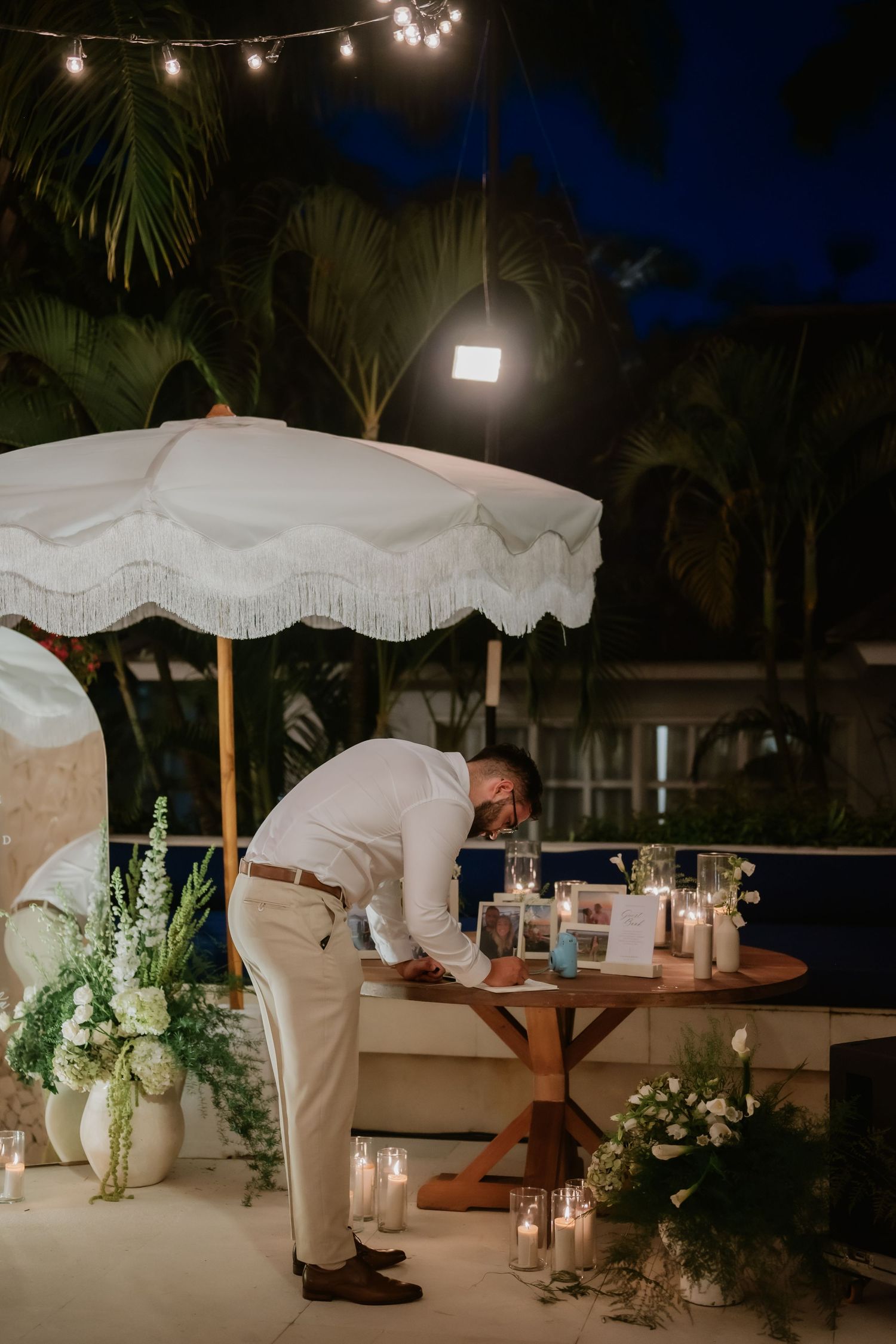 Guest signs guestbook at candlelit table under fringed white umbrella during evening wedding reception.
