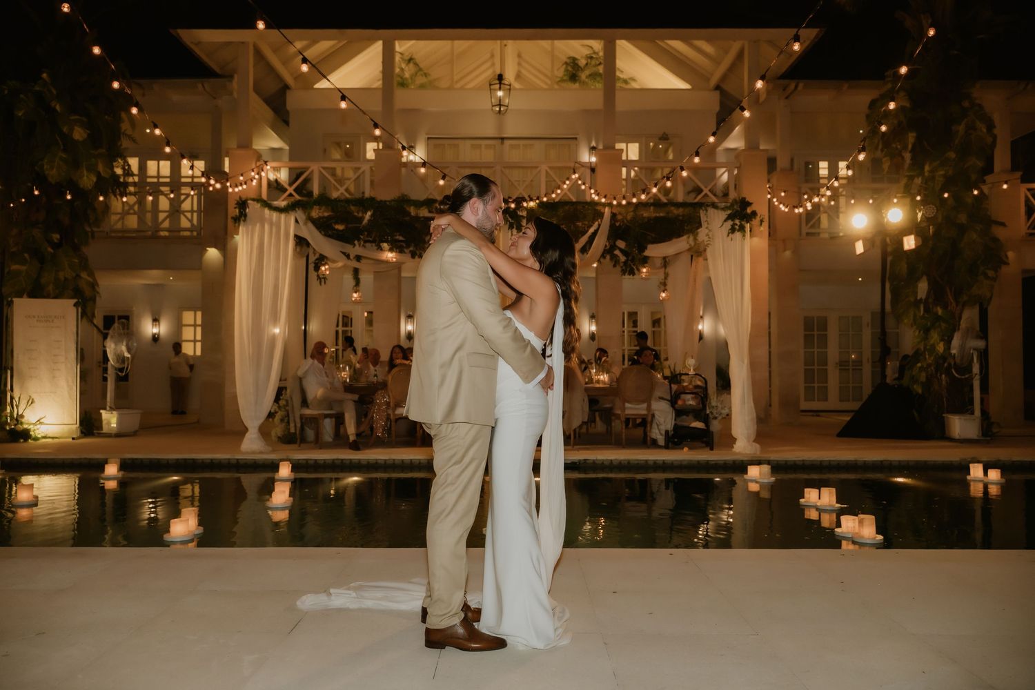 Bride and groom sharing first dance by illuminated pool at romantic tropical wedding reception at night.