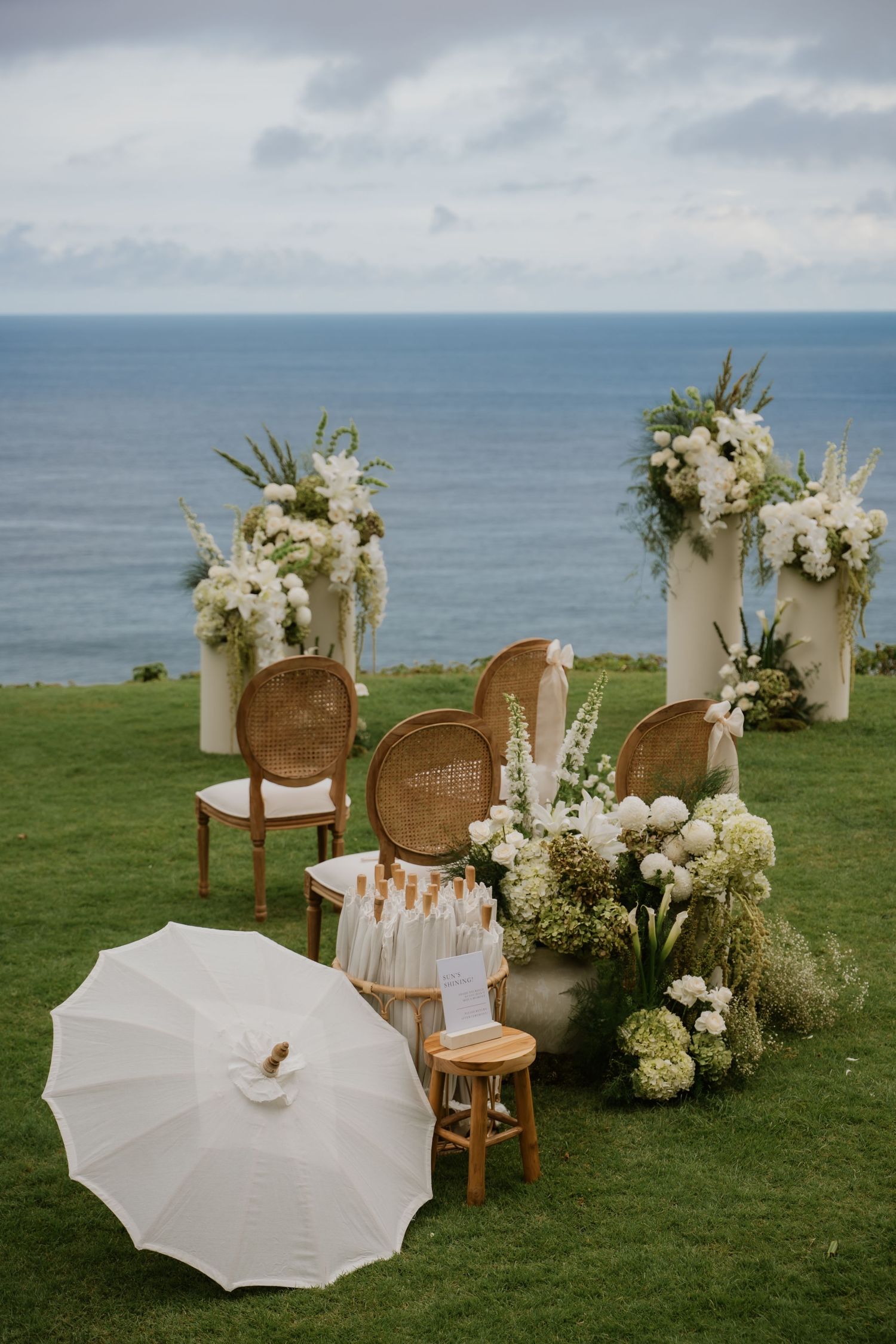 Elegant outdoor wedding ceremony setup with white floral arrangements, rattan chairs, and ocean backdrop in Bali.