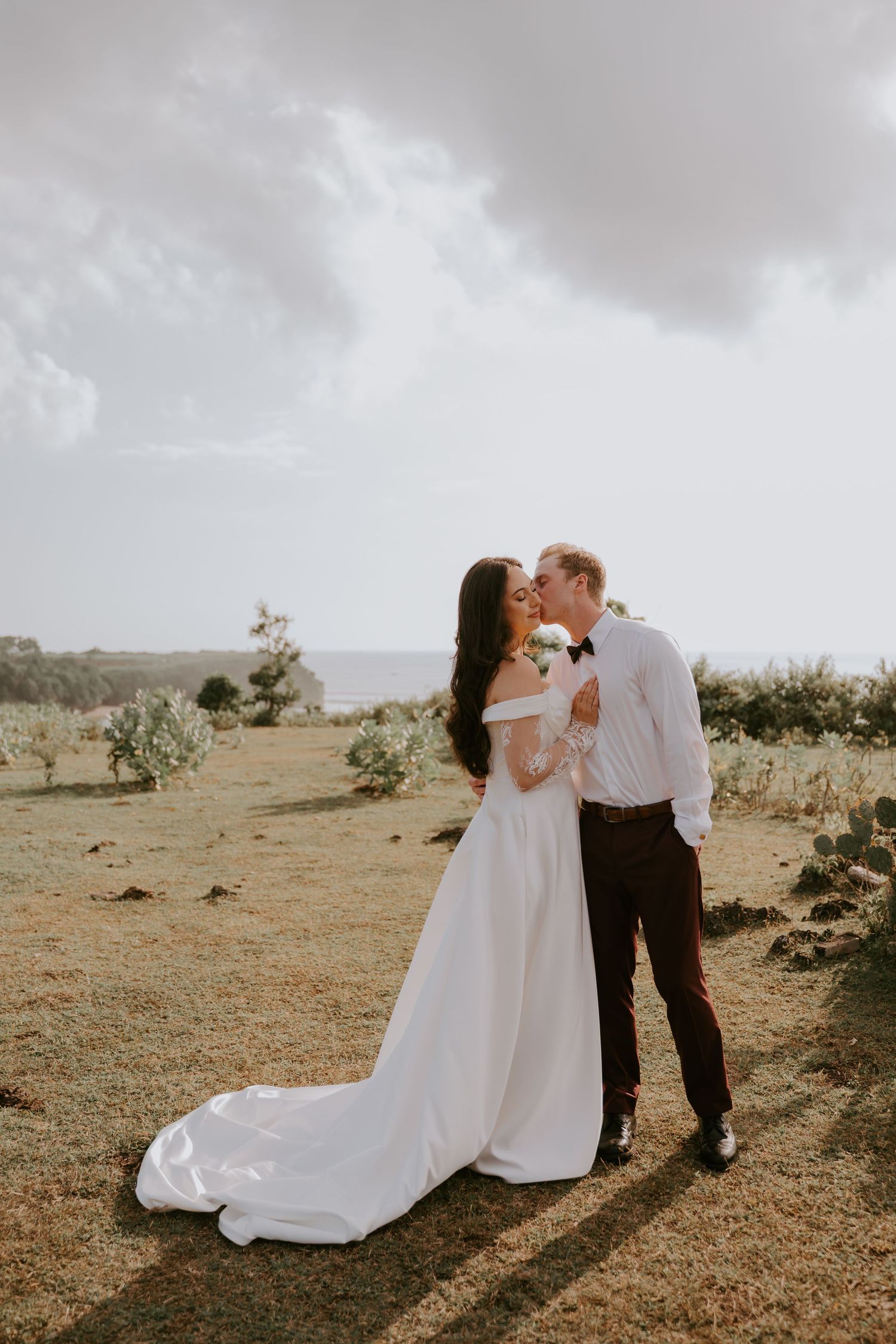 Newlywed couple embracing and kissing in an outdoor coastal setting, bride in elegant off-shoulder white gown with long train.