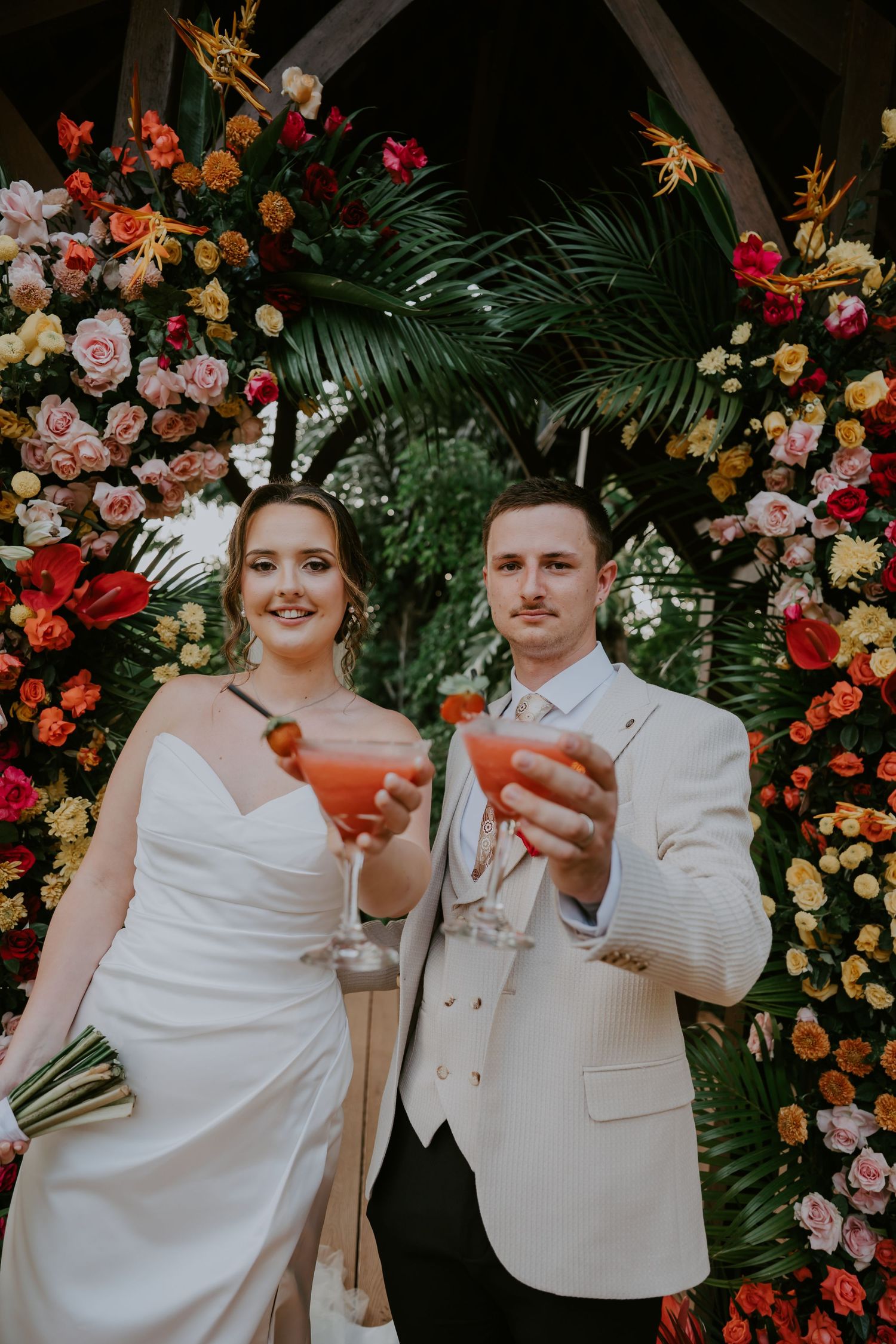 Newlyweds toast with pink cocktails in front of lush colorful floral arch at tropical wedding.