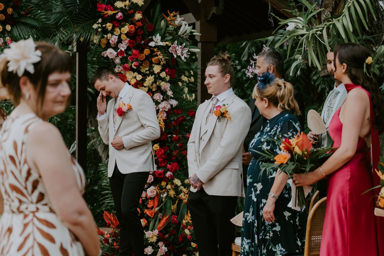 Wedding ceremony guests stand at colorful floral altar as emotional groom wipes tears awaiting bride.