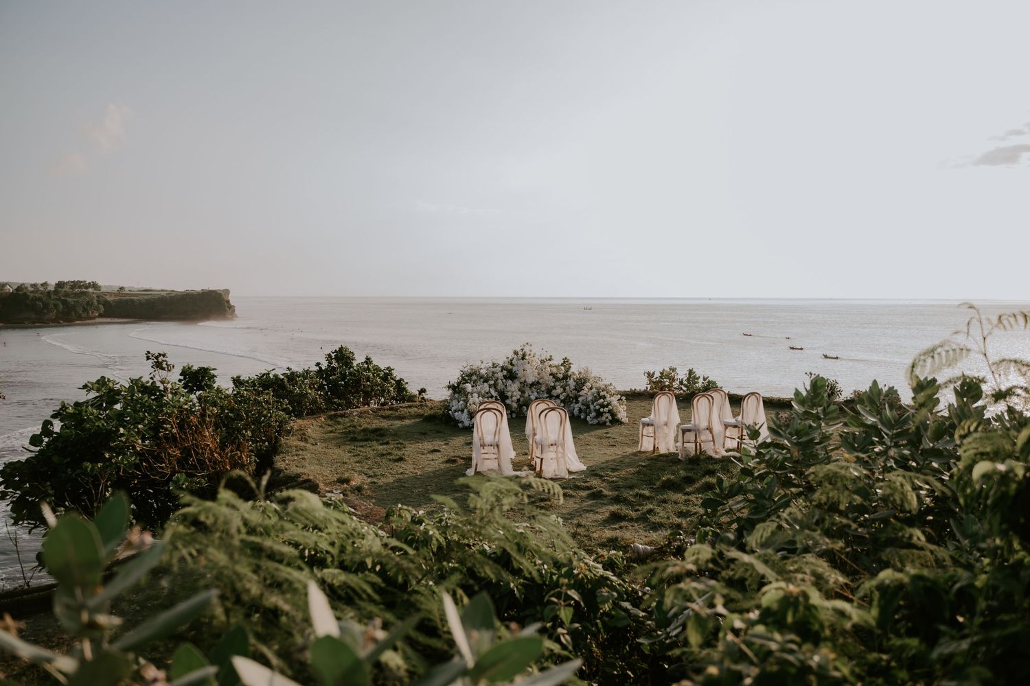 Aerial view of a stunning cliffside wedding ceremony setup with floral arch and draped chairs overlooking the sea.