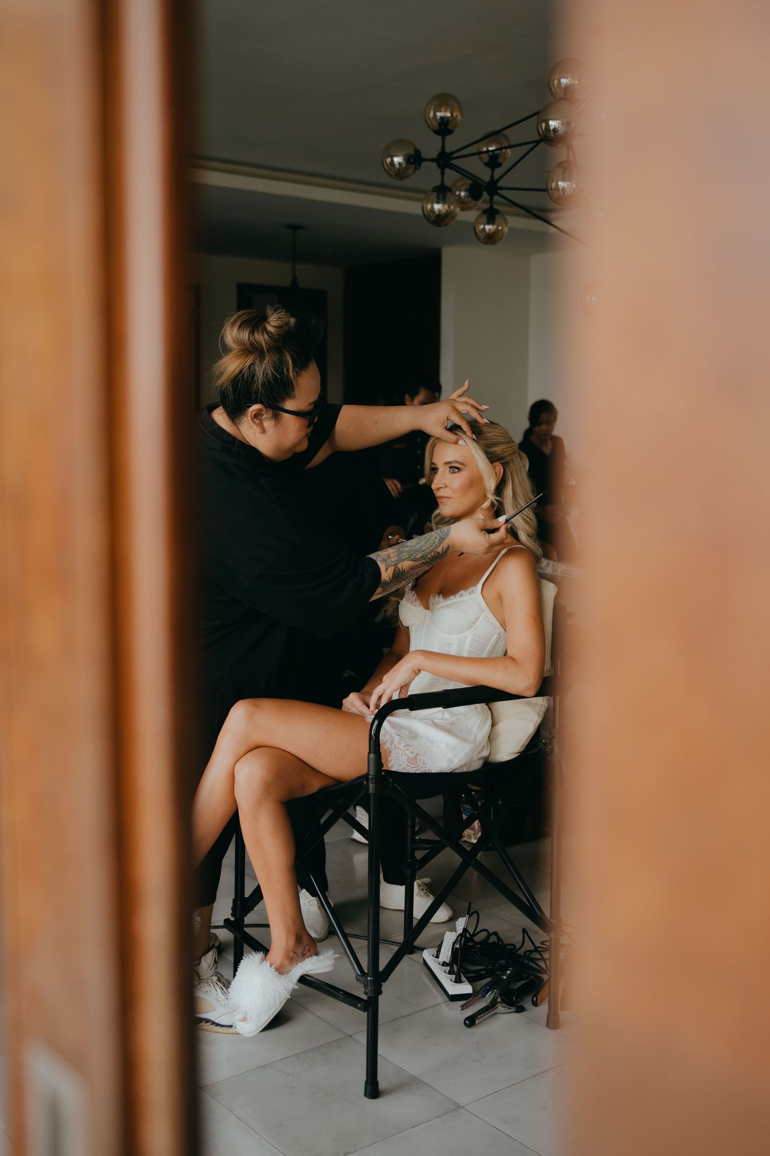 Bride getting hair styled by makeup artist in a white slip dress seated in a salon chair before her wedding.