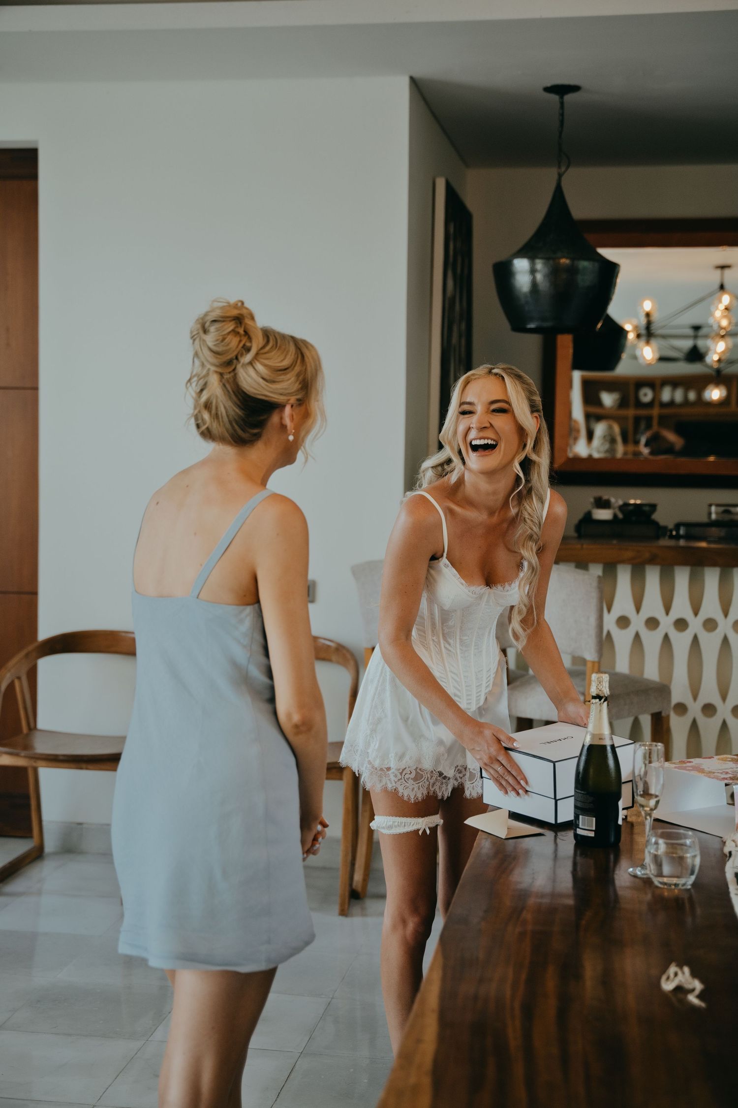 Bride in white lace lingerie laughing joyfully with bridesmaid during wedding morning preparations.