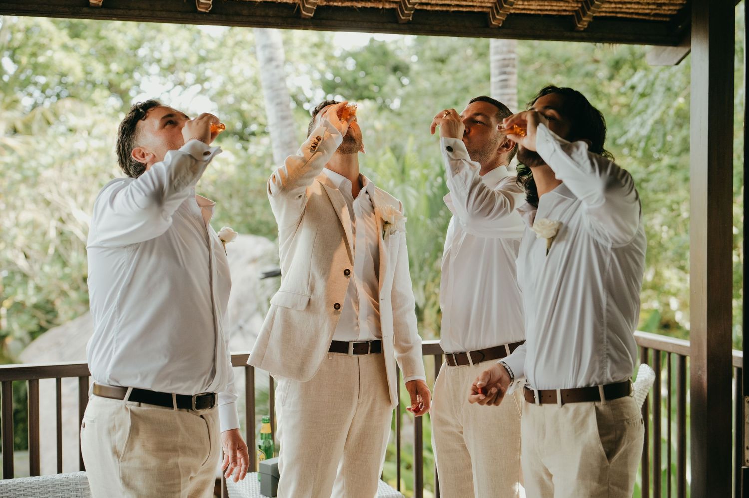 Groomsmen in white shirts doing shots together on outdoor porch before wedding ceremony.