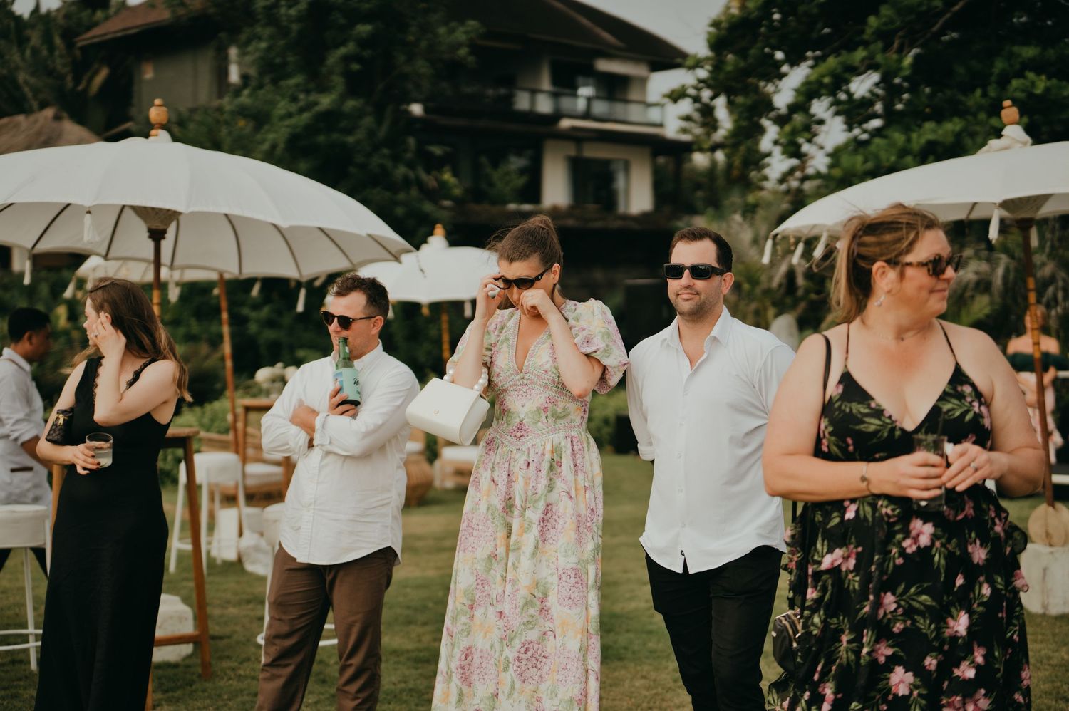 Wedding guests in summer attire socializing outdoors under white parasols at a garden reception.