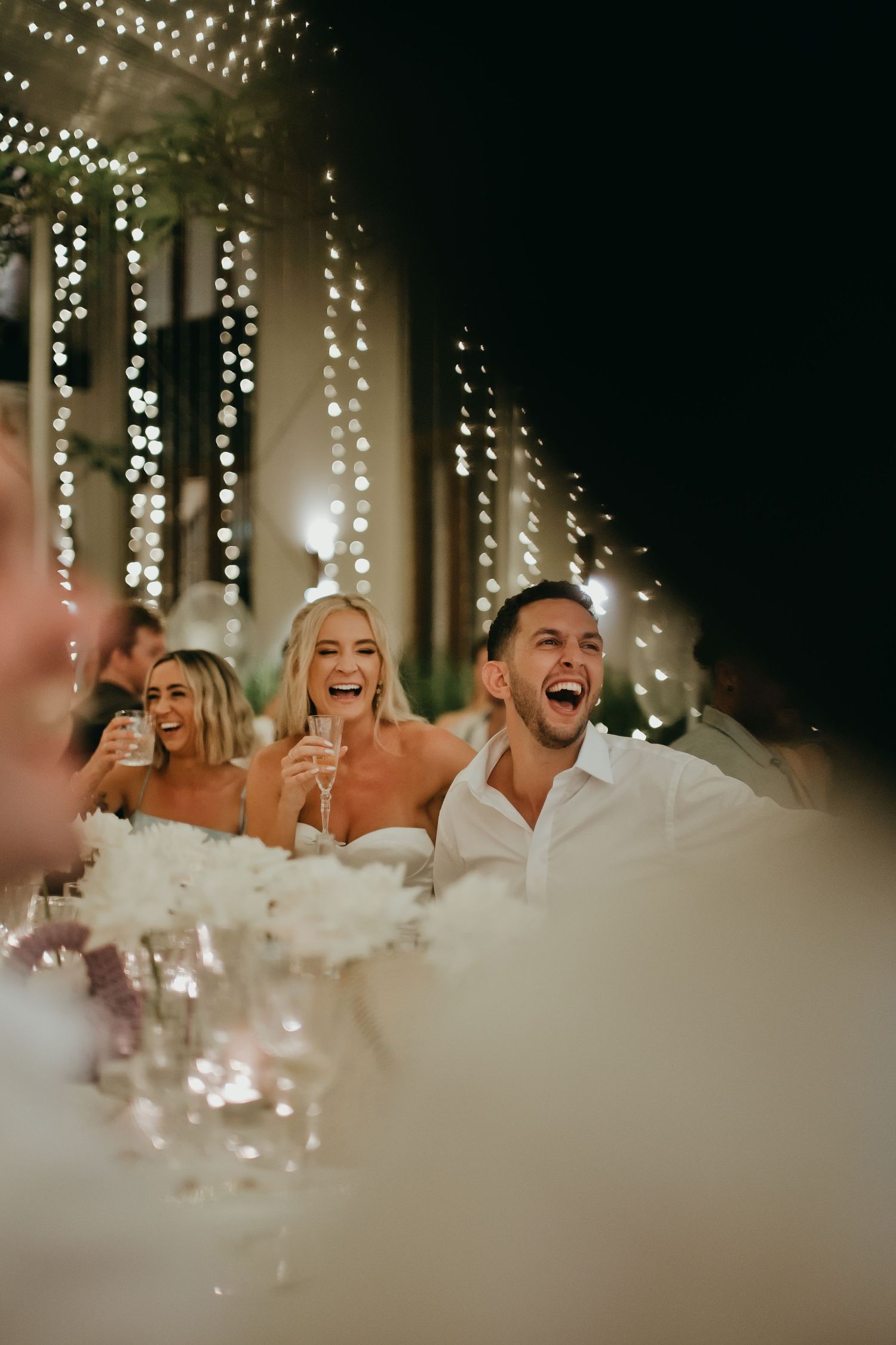 Laughing bride and groom enjoy speeches at candlelit reception table adorned with white flowers and fairy lights.