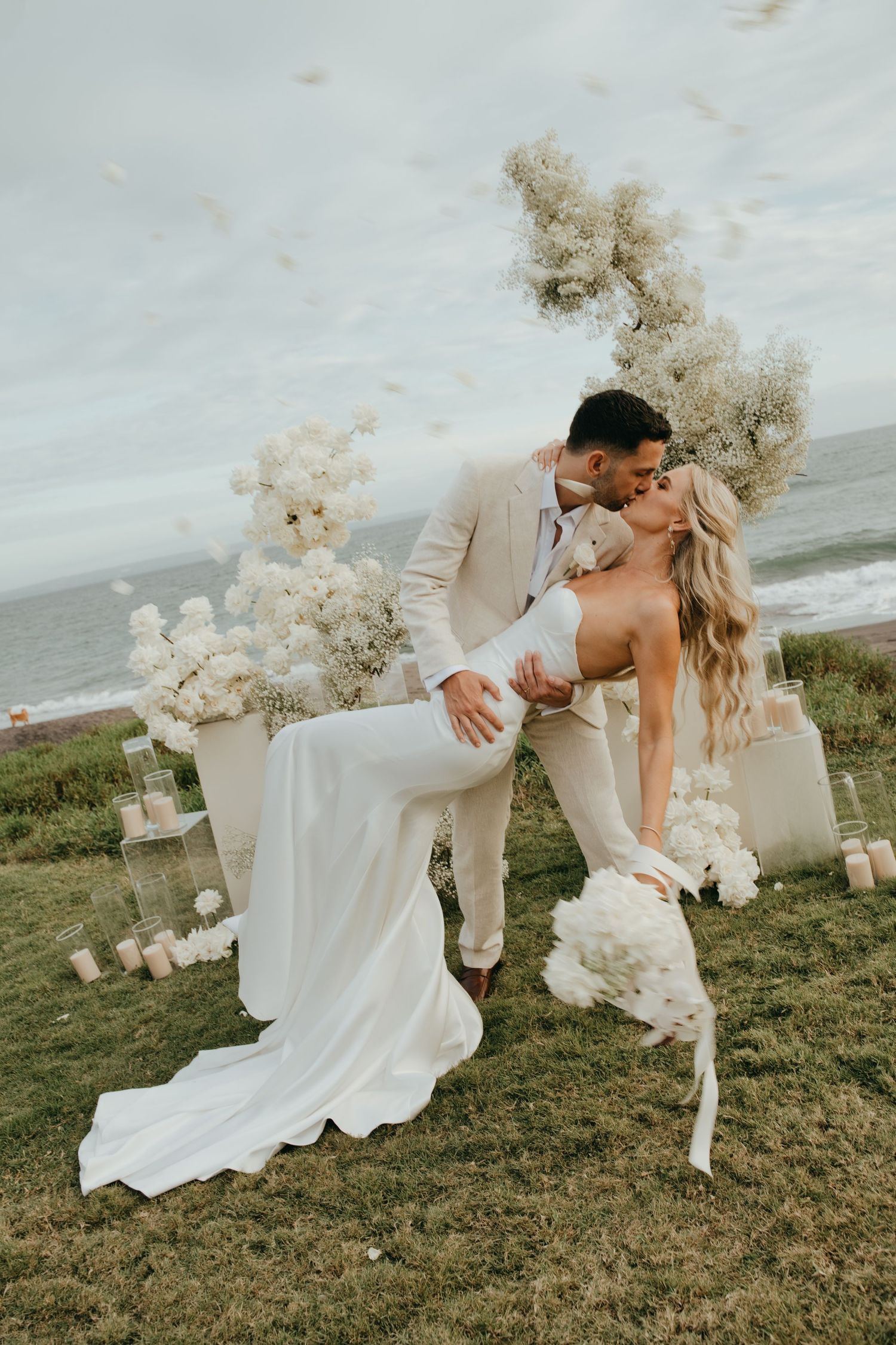 Bride and groom share a romantic kiss surrounded by white floral arrangements and candles at an oceanside wedding ceremony.