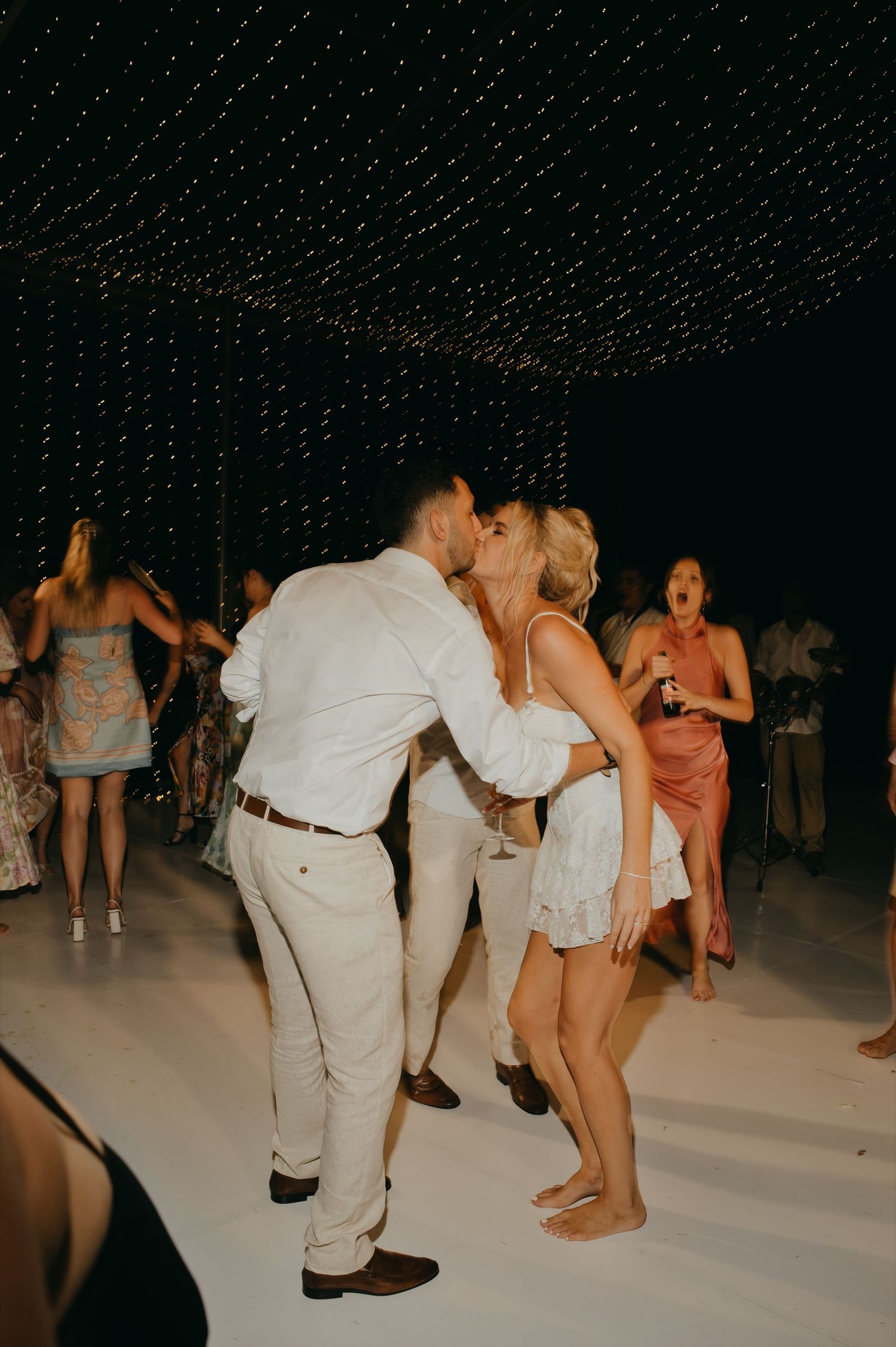Bride and groom sharing a kiss on the dance floor while guests celebrate around them at night.