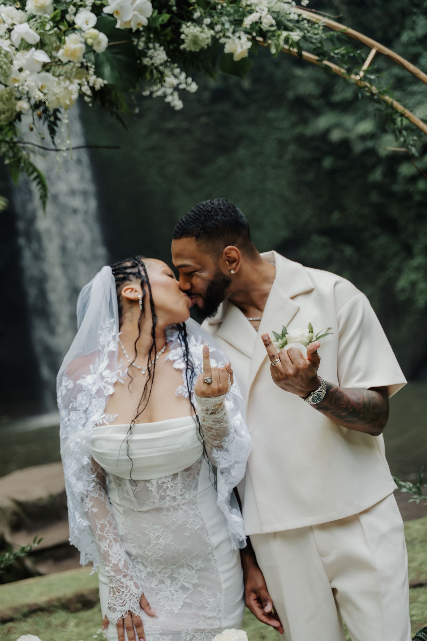 Newlyweds share their first kiss beneath a white floral arch with a tropical waterfall in background.