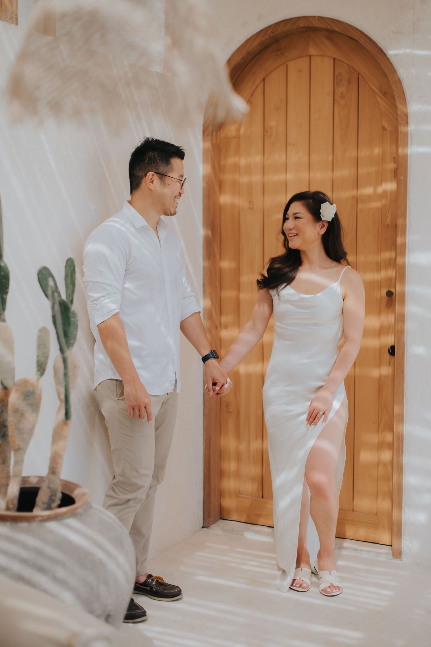 Couple in white outfits holding hands before an arched wooden door, surrounded by cacti in a bright, airy setting.