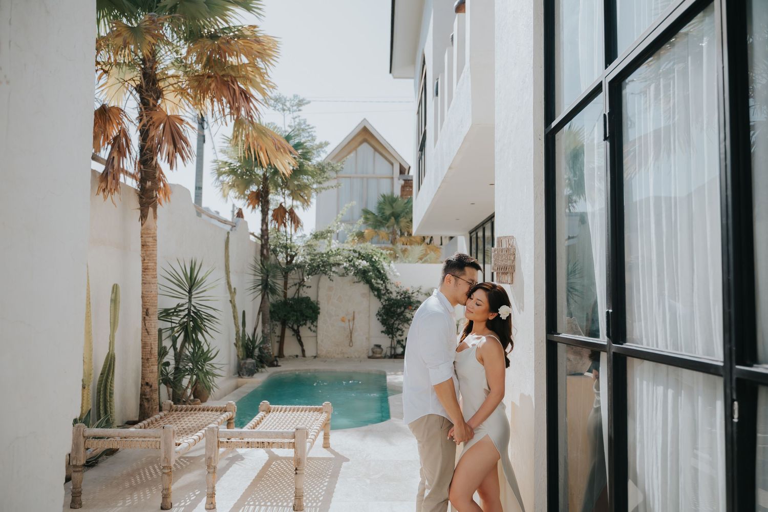 Couple sharing a romantic moment by a pool in a tropical villa courtyard with palm trees and lush greenery.