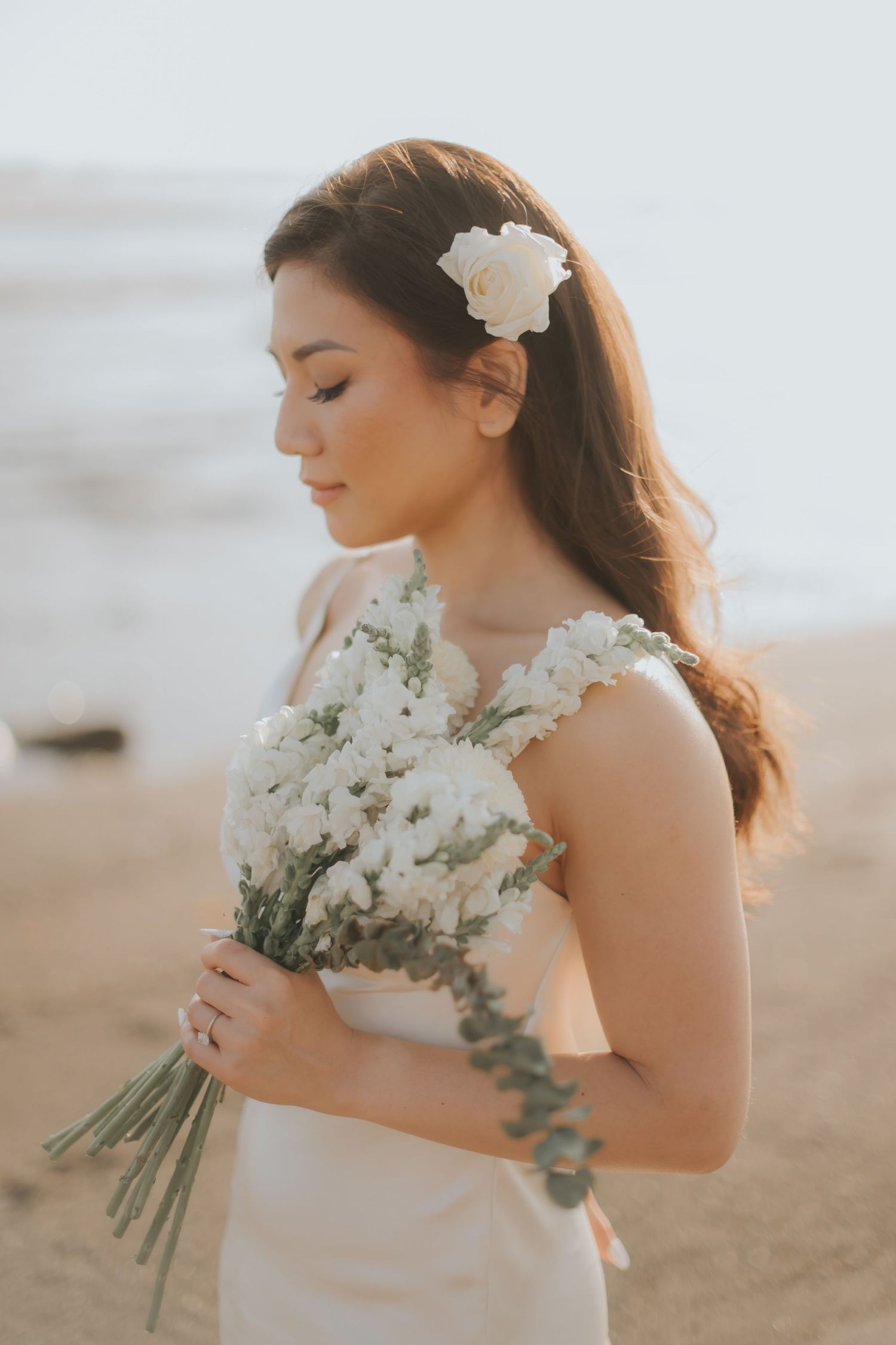 A bride in a white dress holds a bouquet of white flowers on a beach, with a floral hair accessory.