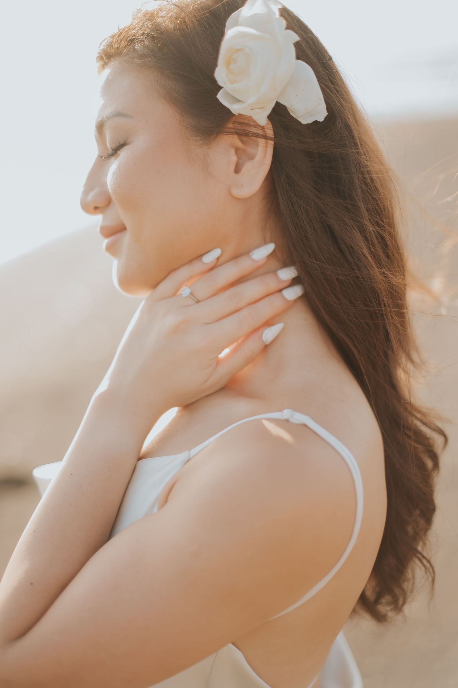A serene woman with a white flower in her hair, eyes closed, wearing a white dress, gently touching her neck outdoors.