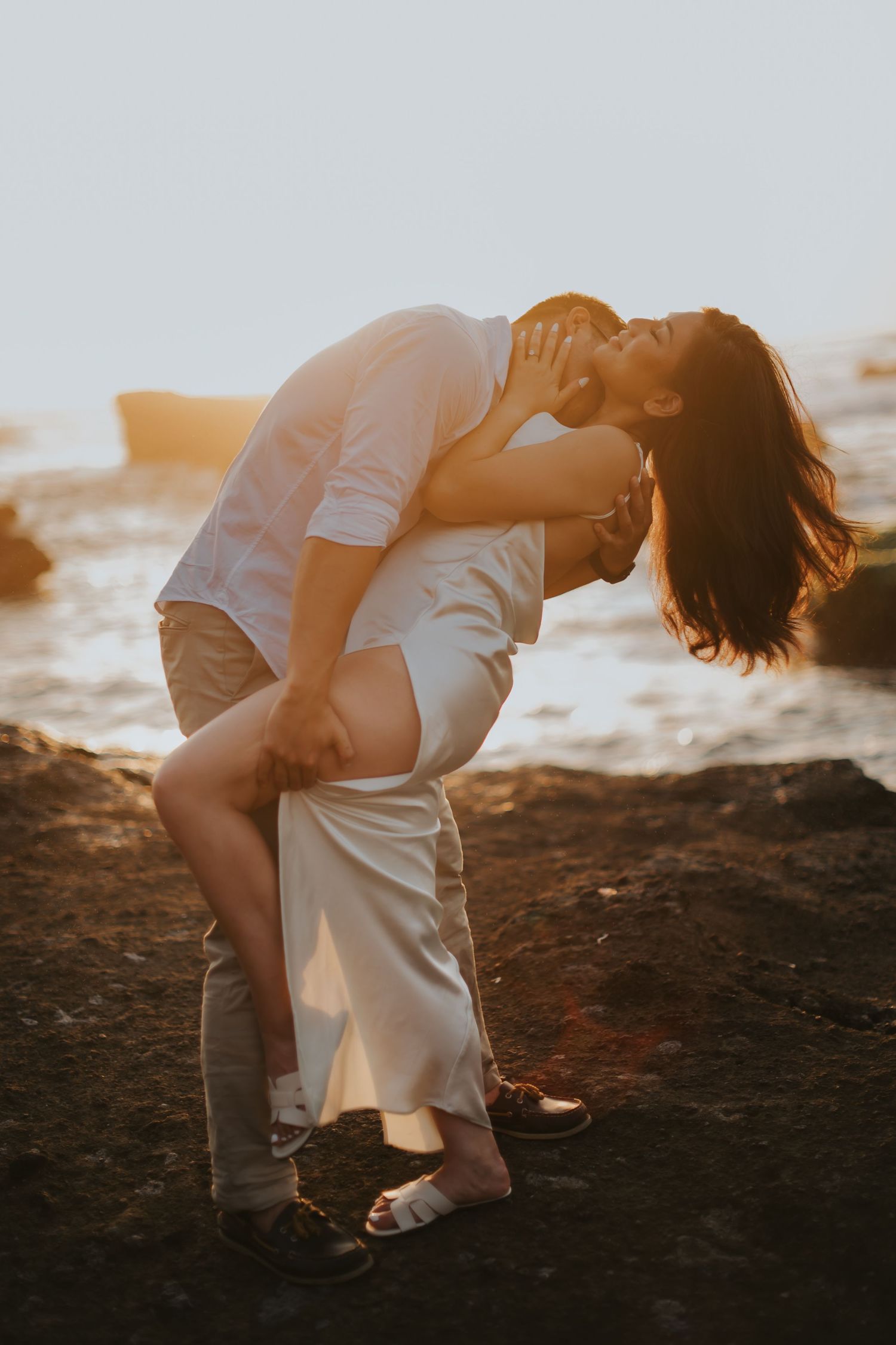 Couple sharing a romantic kiss at golden hour on rocky beach, man lifting woman in white dress at sunset.