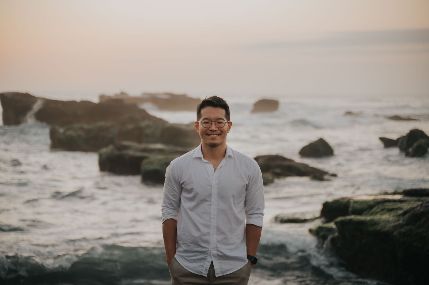 Smiling young Asian man in white shirt standing on rocky ocean shore at sunset.