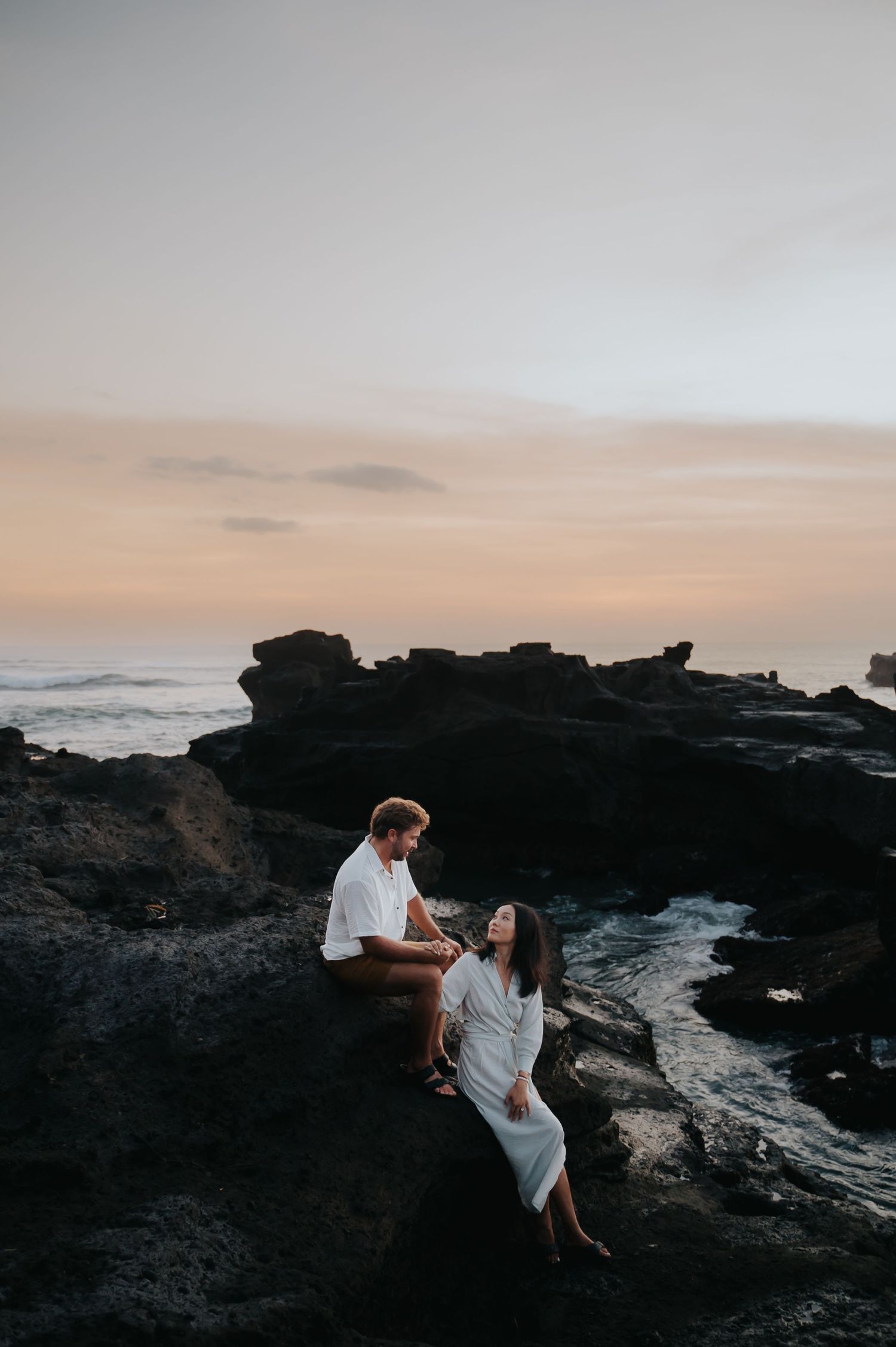 A couple in white outfits sits together on dark volcanic rocks at sunset along a dramatic coastline.