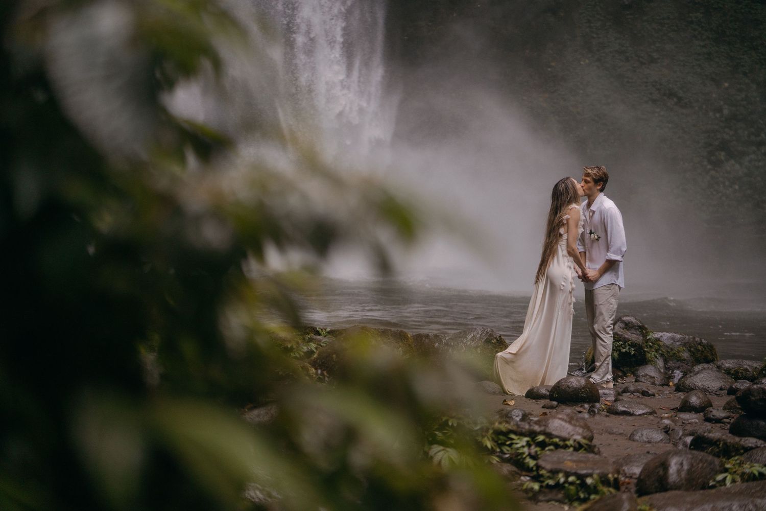 A couple in wedding attire embraces on misty rocky terrain with soft lighting and blurred foliage in the foreground.