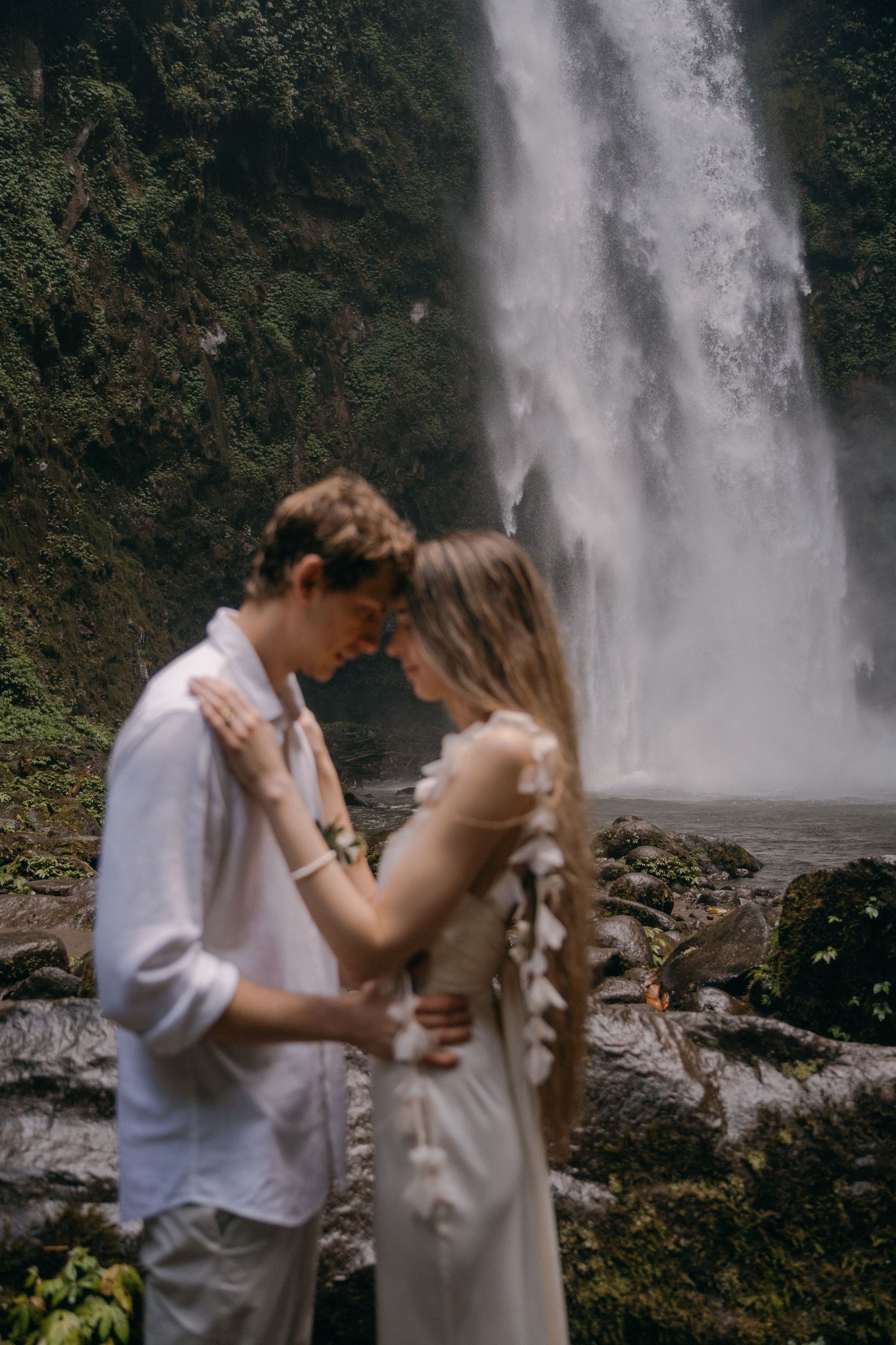A couple shares an intimate moment in front of a majestic waterfall in a lush forest setting.