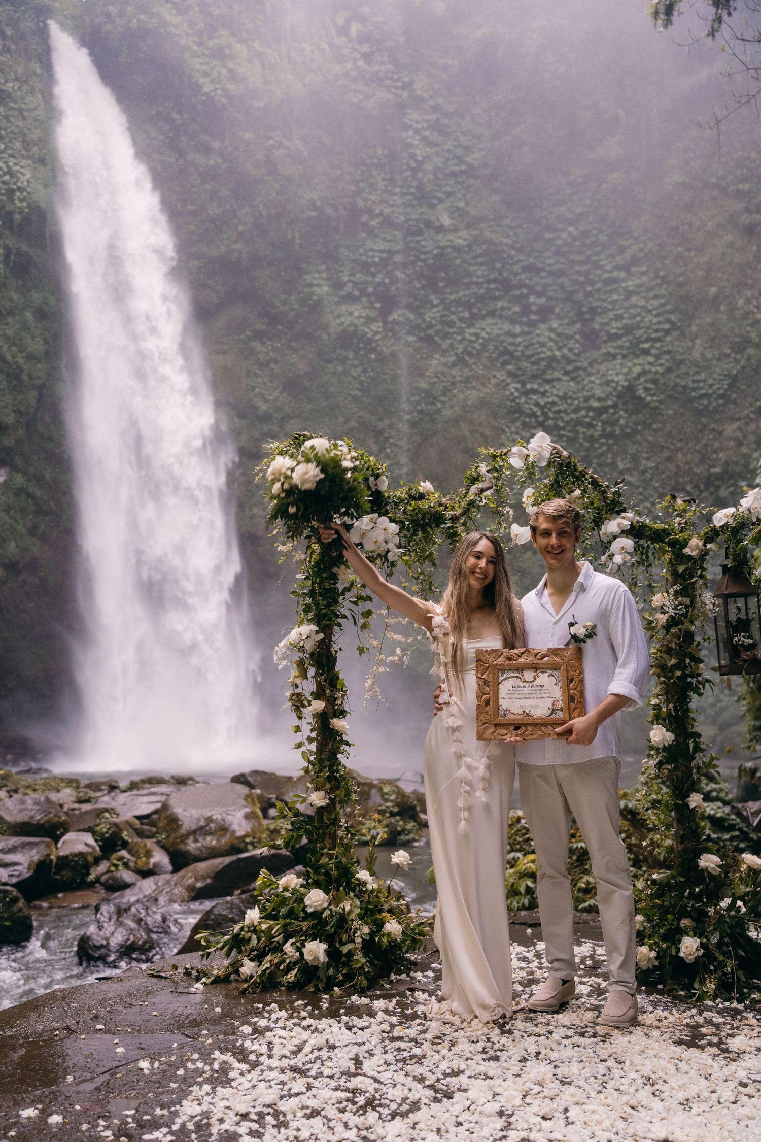 A romantic outdoor wedding ceremony setup with floral arch and waterfall backdrop in a misty forest landscape.
