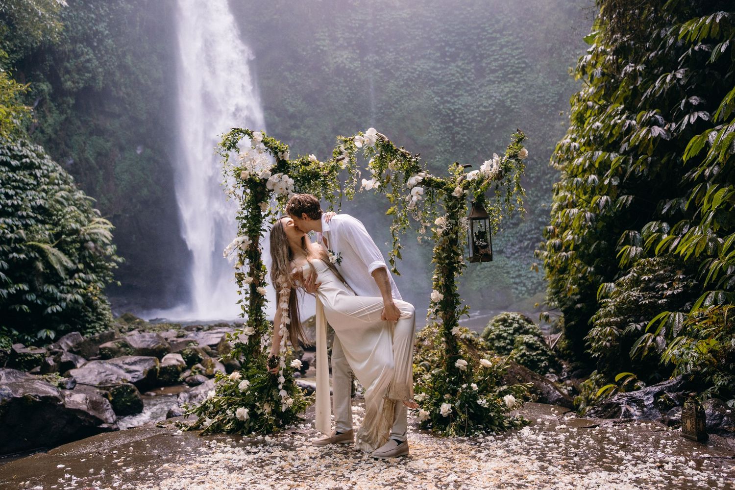 A couple in light-colored outfits poses together near a scenic tropical waterfall in a lush forest setting.