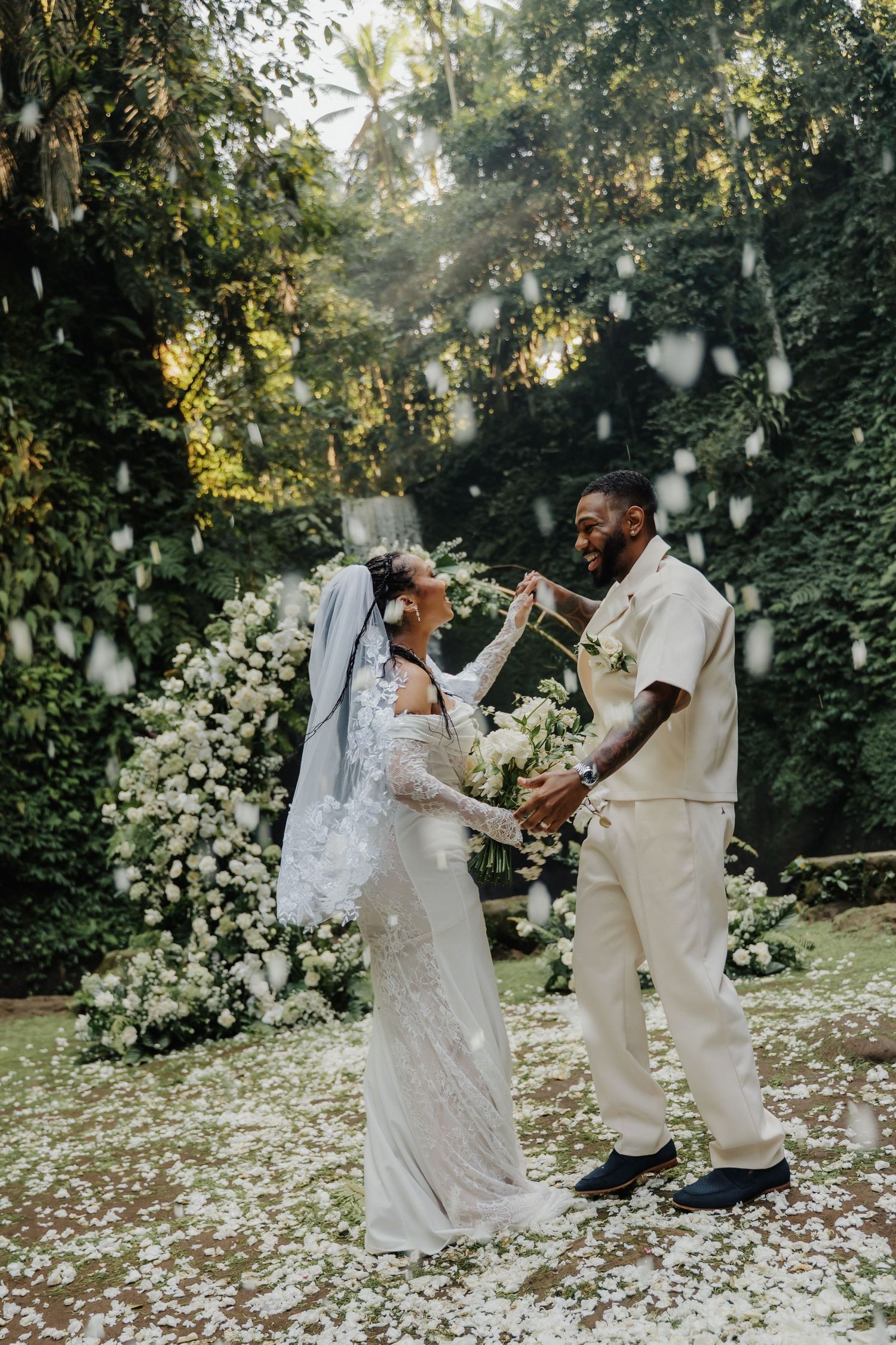 Couple dancing joyfully at outdoor wedding ceremony with white floral arch and falling petals in lush garden setting.