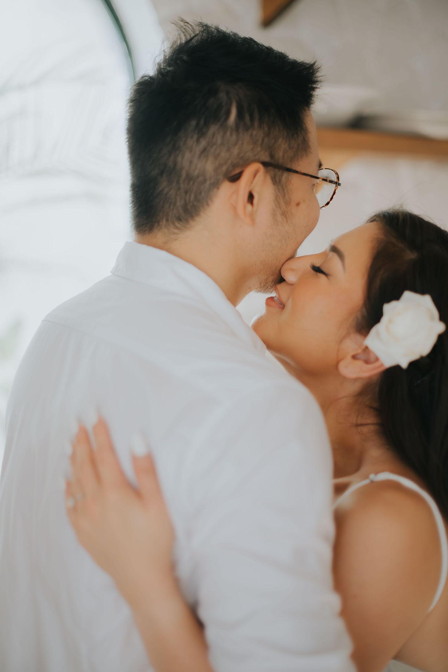 A couple shares a tender kiss, dressed in white, with a flower in the woman's hair.