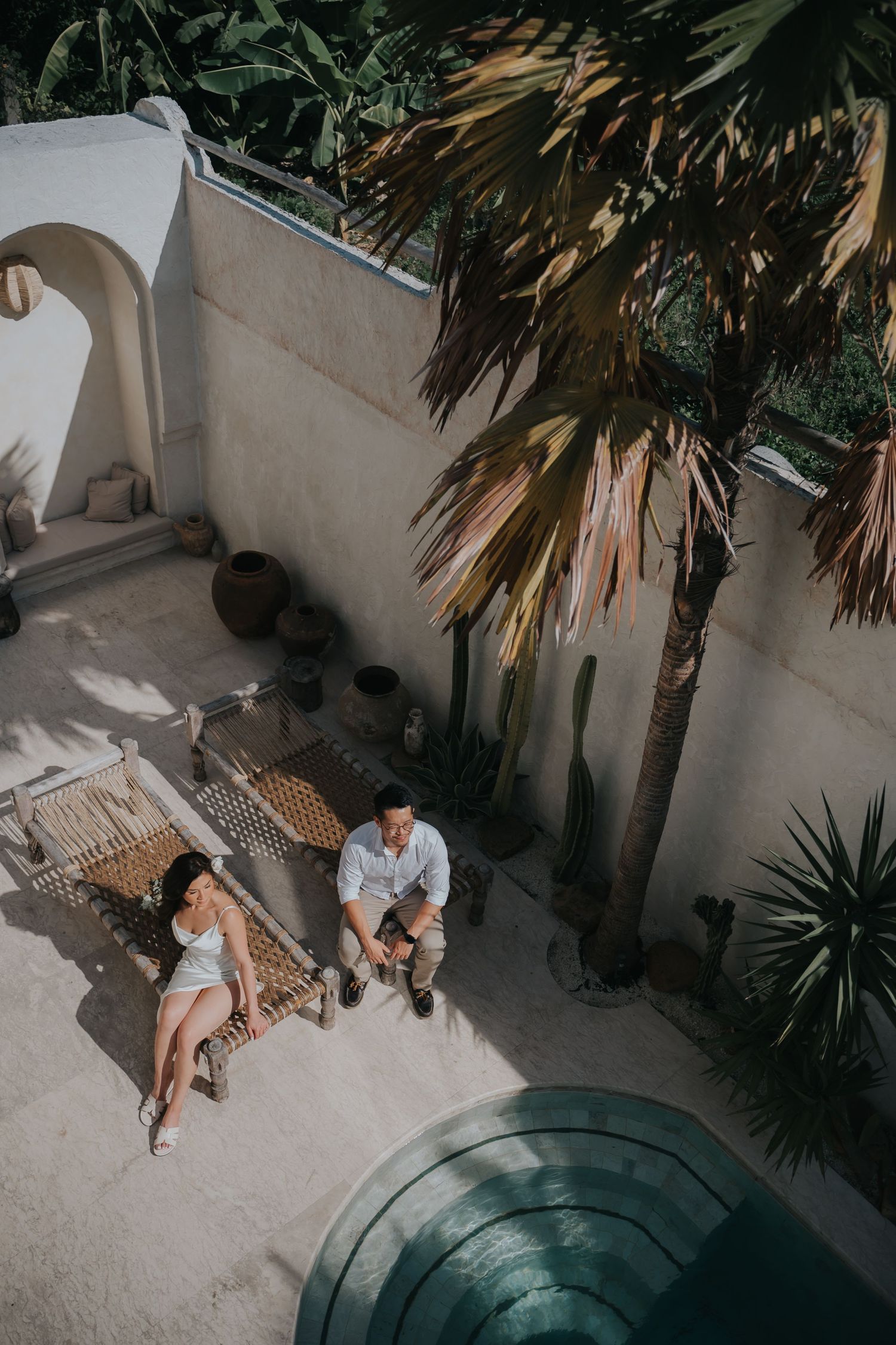 Aerial view of a couple relaxing on a wooden daybed in a tropical courtyard with palm trees and a pool.