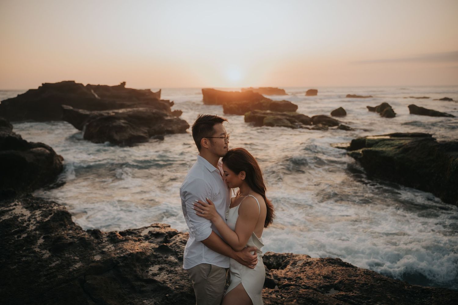Couple embracing at sunset on rocky ocean shore with waves crashing behind them.