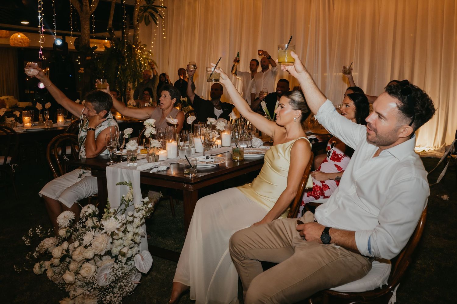 Wedding guests raise glasses in a toast at candlelit reception dinner table with floral arrangements.