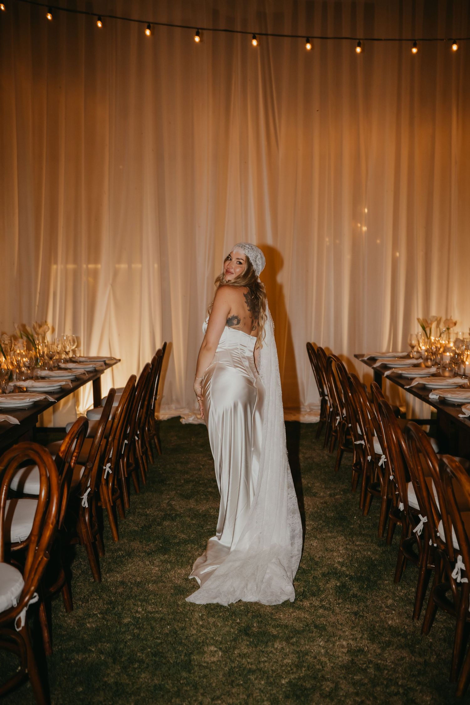 Bride in backless white gown and veil stands between elegant reception tables with warm draping.