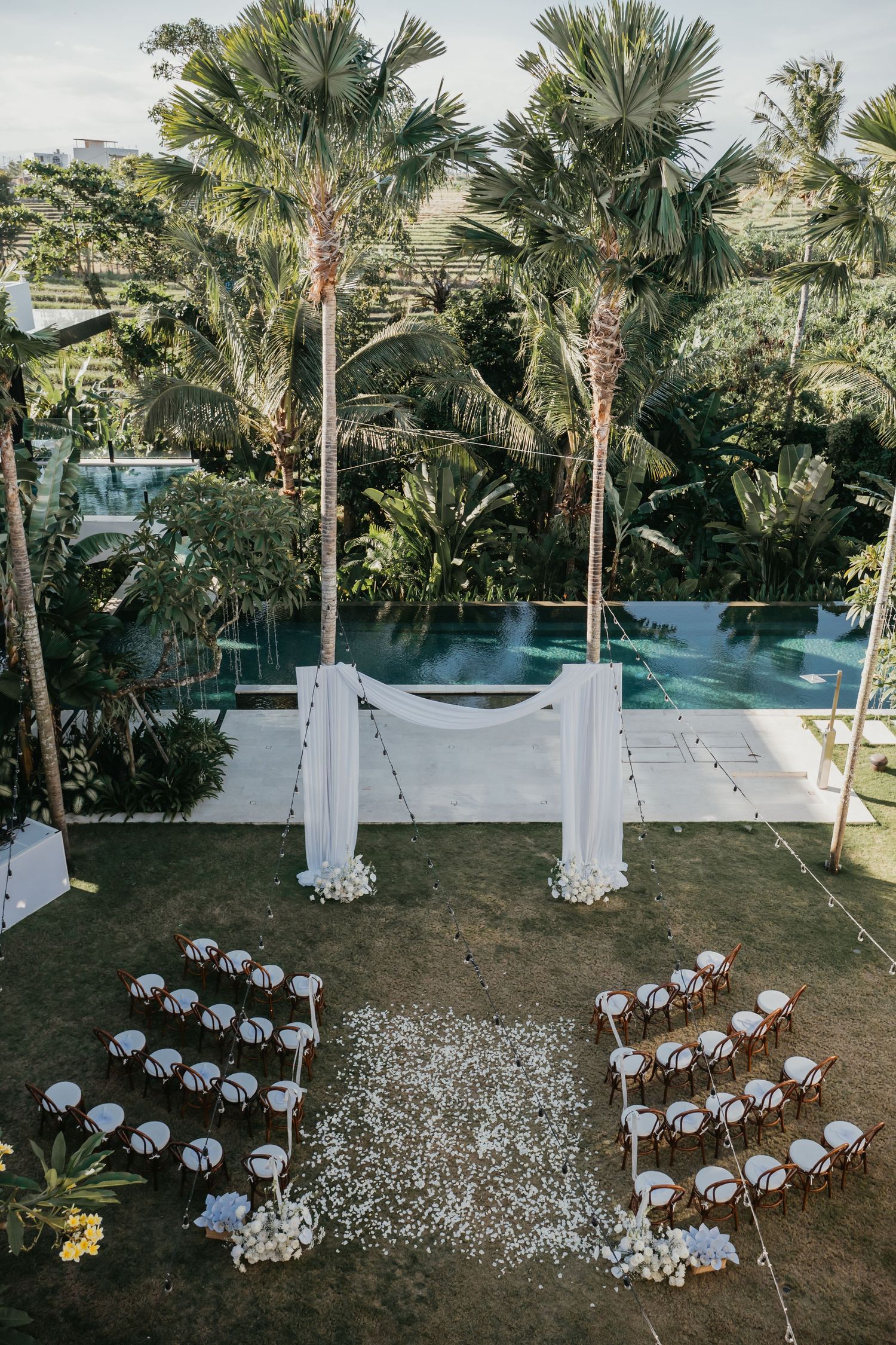 Aerial view of outdoor wedding ceremony with white arch, petal aisle, and pool surrounded by palm trees.