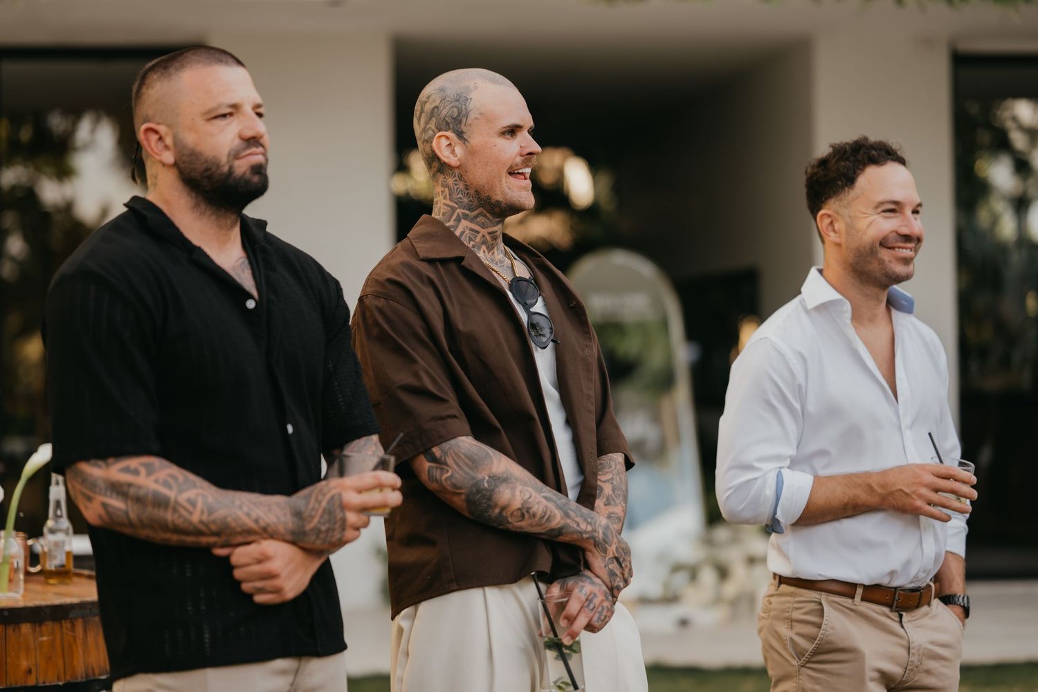 Three tattooed groomsmen smiling and watching outdoor wedding ceremony proceedings.