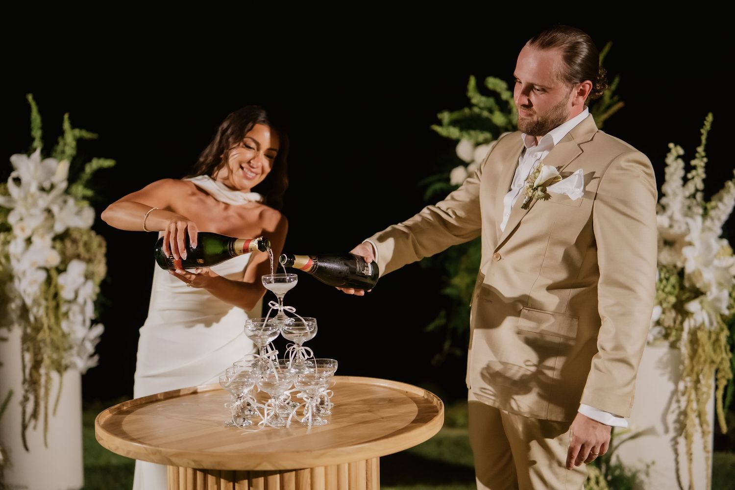Bride and groom pour champagne tower together at outdoor wedding reception with white floral arrangements.