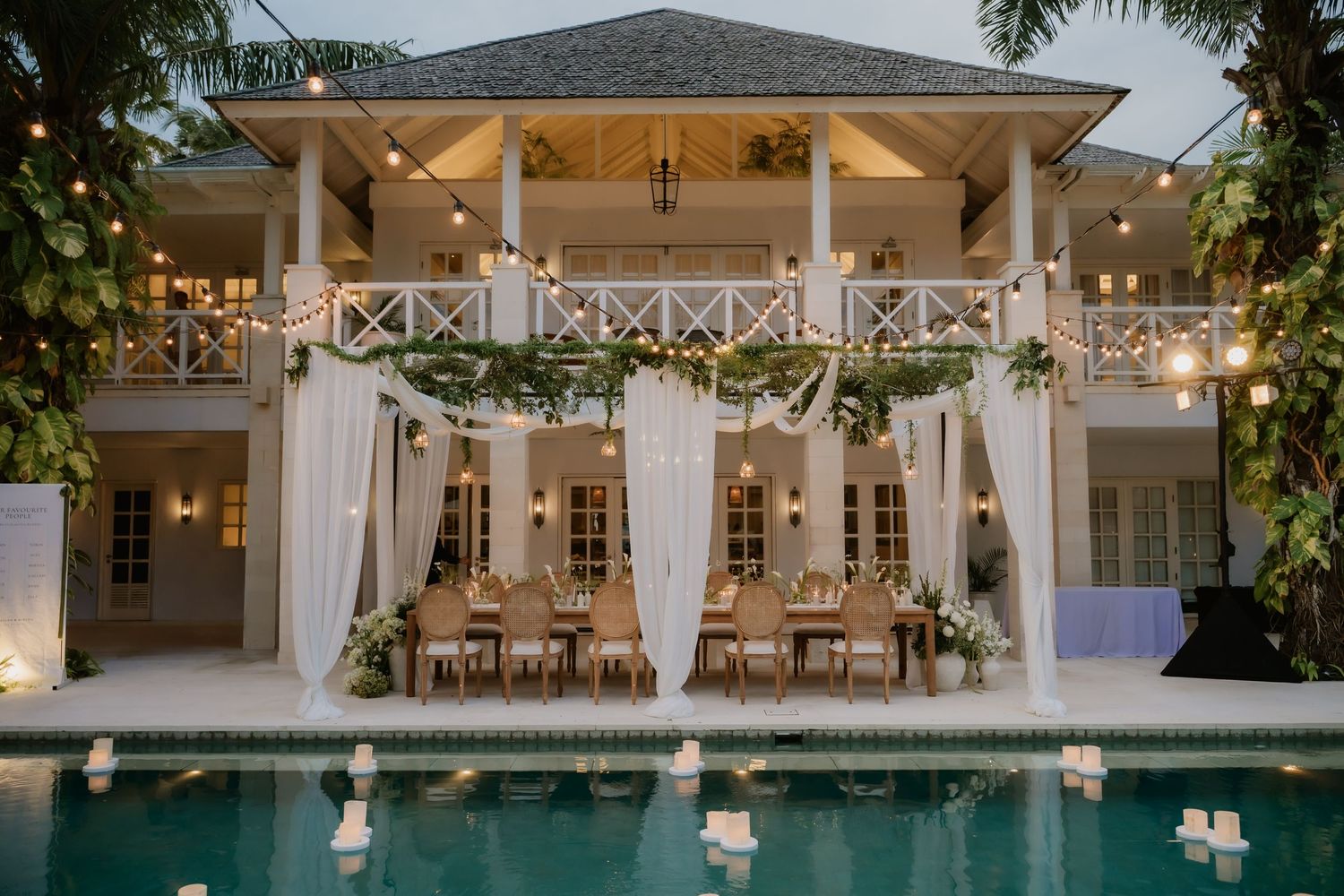 Elegant tropical wedding reception setup beside pool with string lights and draped white curtains at dusk.