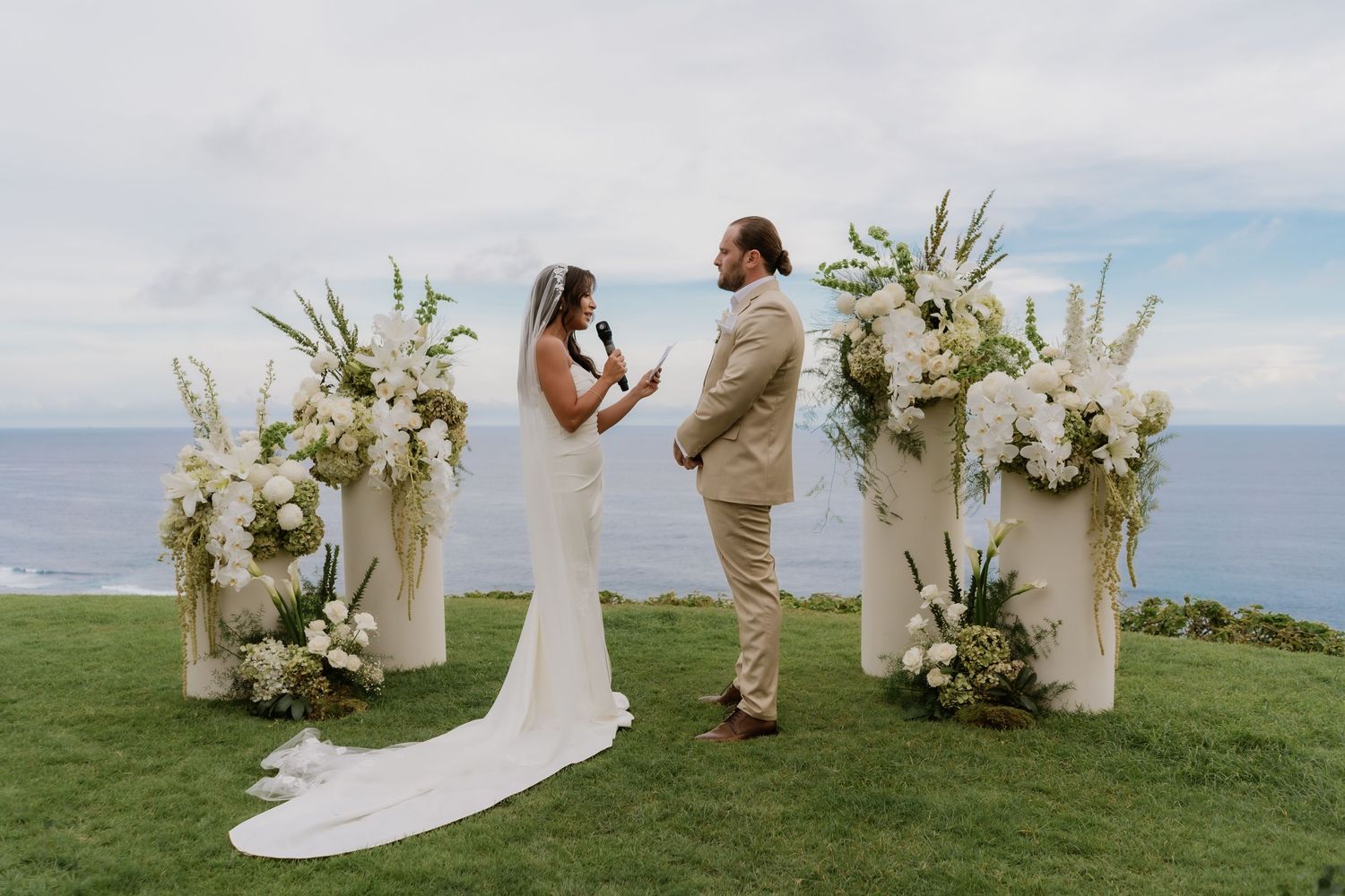 Bride and groom stand at a cliffside wedding altar adorned with tall white floral arrangements overlooking the ocean.
