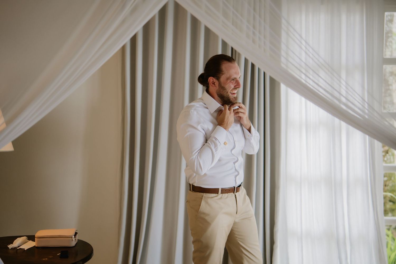 Groom in white shirt and tan pants adjusting his tie while smiling near sheer curtains in a sunlit room.