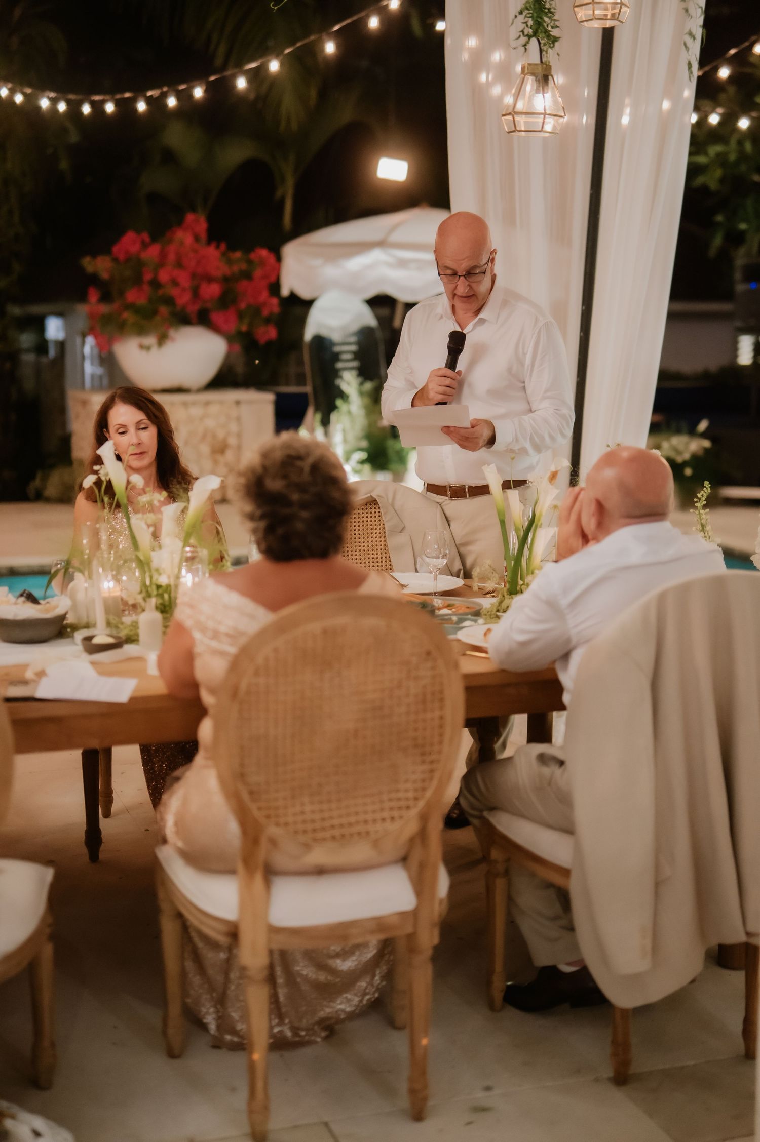 Bald man delivers speech at outdoor wedding reception while guests listen at decorated table.