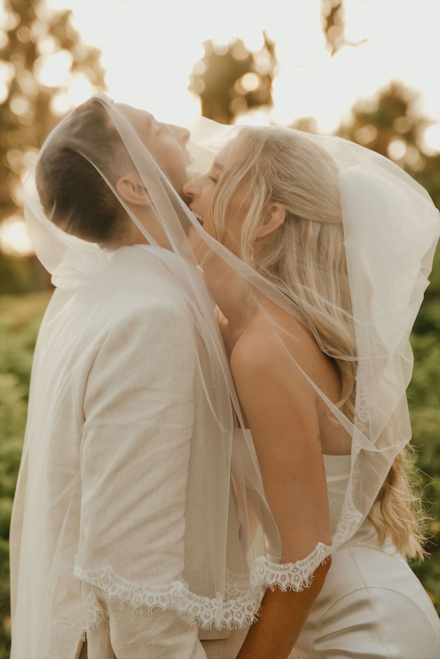 Romantic golden hour portrait of bride and groom sharing a kiss beneath a delicate lace-trimmed veil.