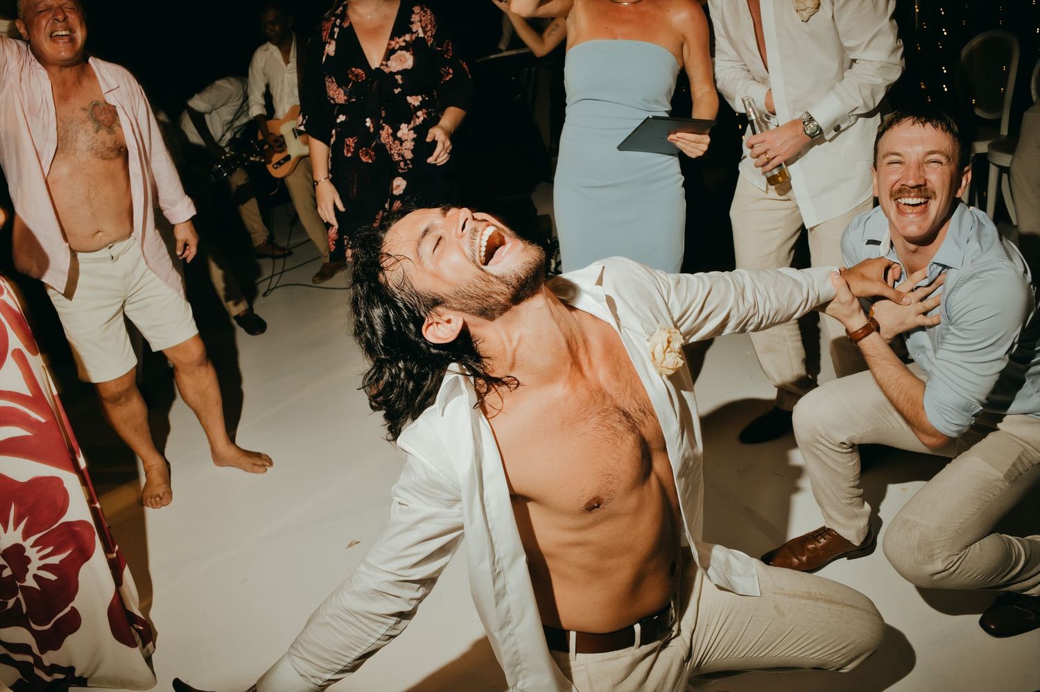 Energetic wedding guest dancing shirtless on the dance floor surrounded by laughing celebrating guests.