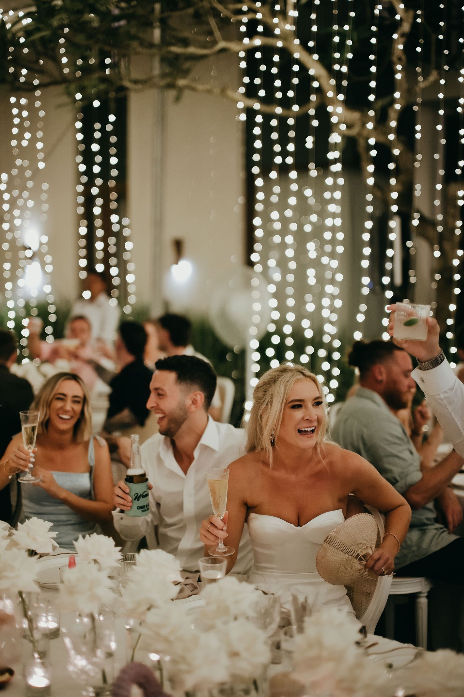 Bride and groom laugh joyfully at reception table surrounded by white floral arrangements and twinkling fairy lights.