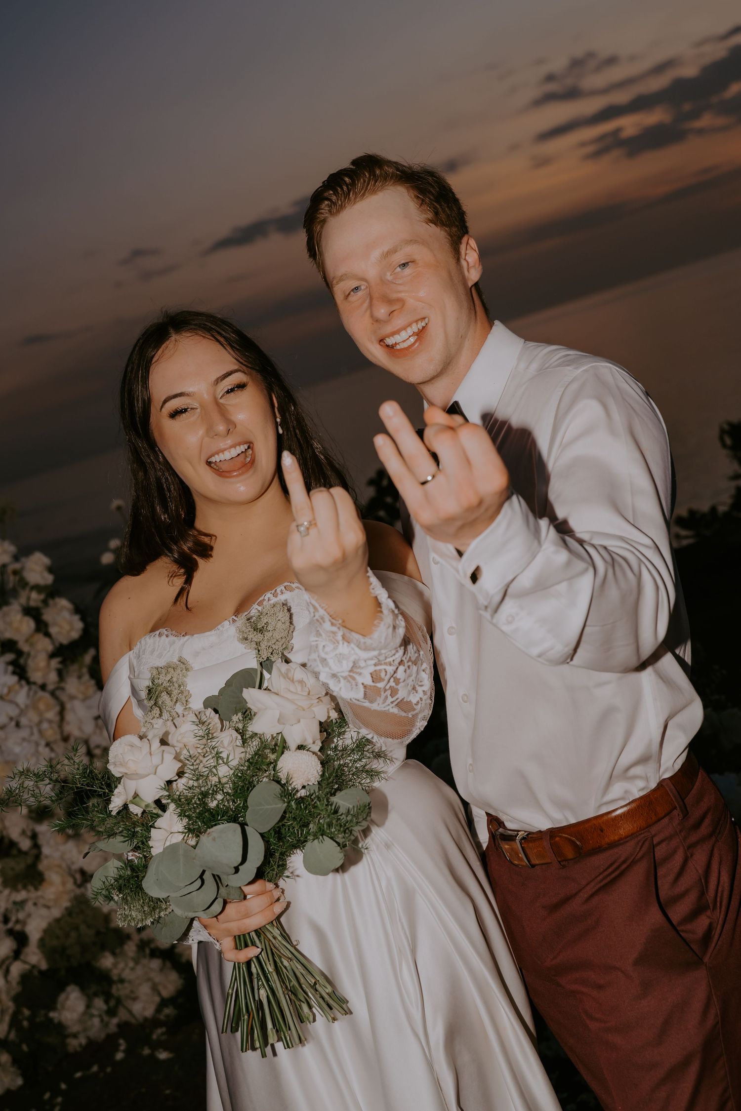 Joyful newlyweds show off wedding rings while bride holds white and green bouquet at sunset reception.