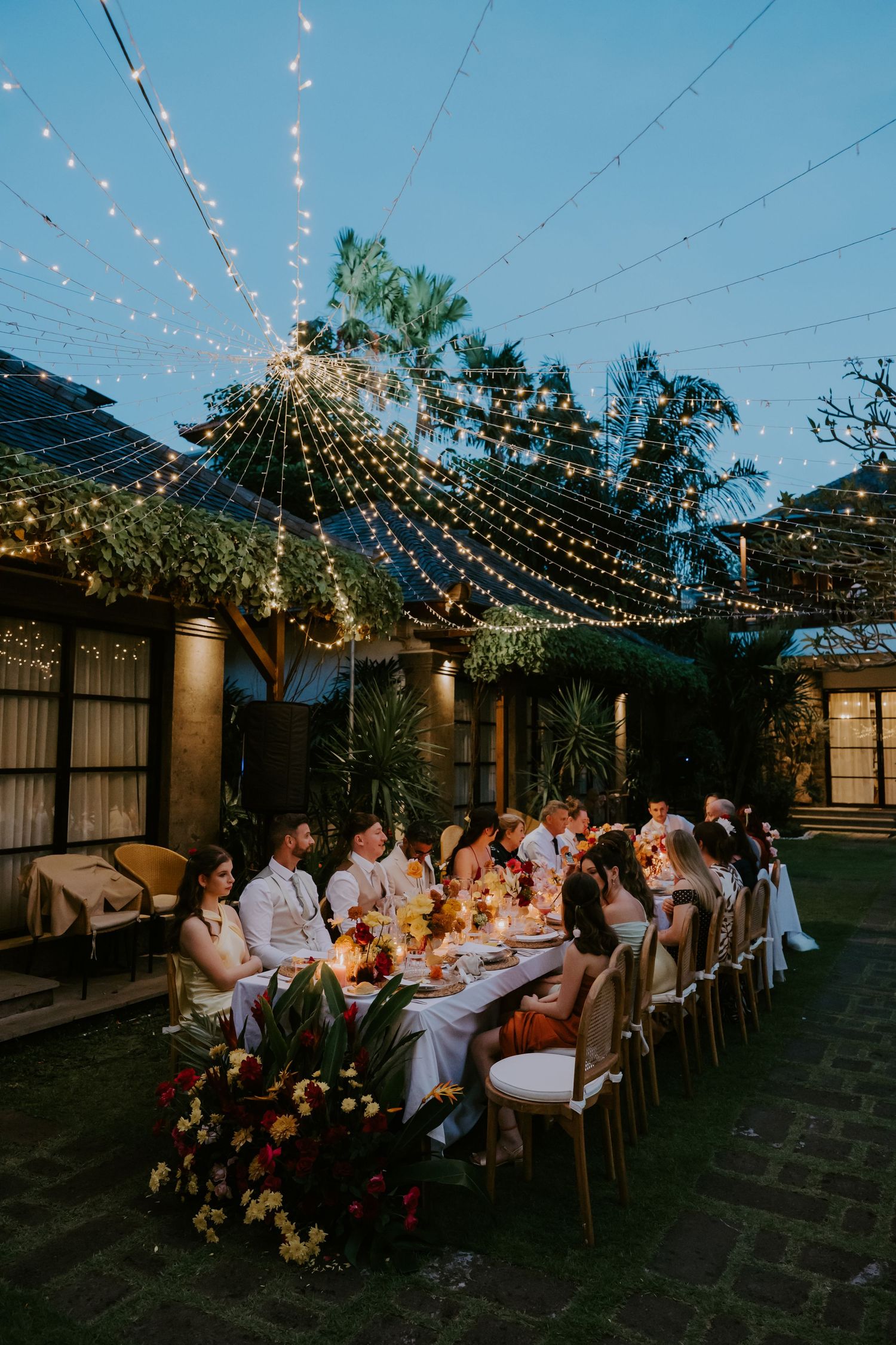 Wedding guests dine at an elegant outdoor reception table illuminated by string lights at twilight.