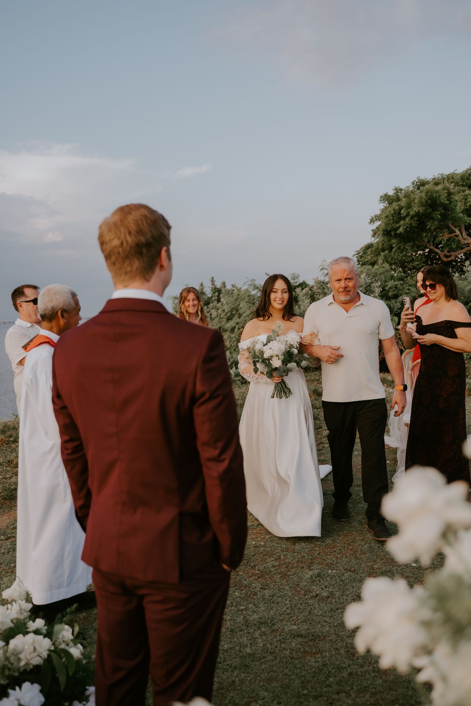Bride in white off-shoulder gown walks down the aisle with her father toward the groom during an outdoor sunset wedding ceremony.
