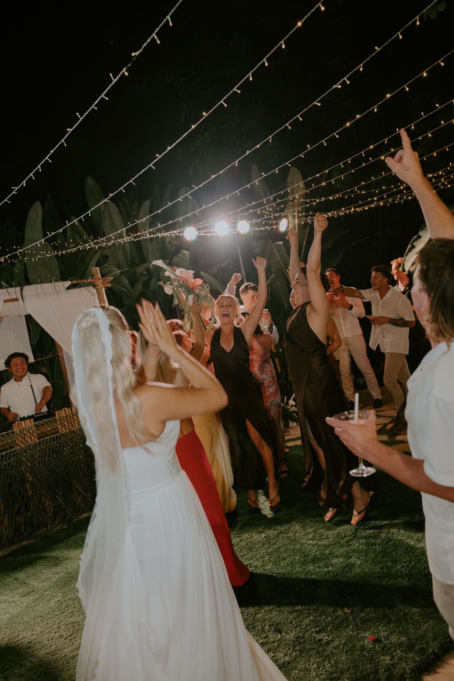 Bride celebrated by joyful wedding guests dancing under string lights at outdoor night reception.